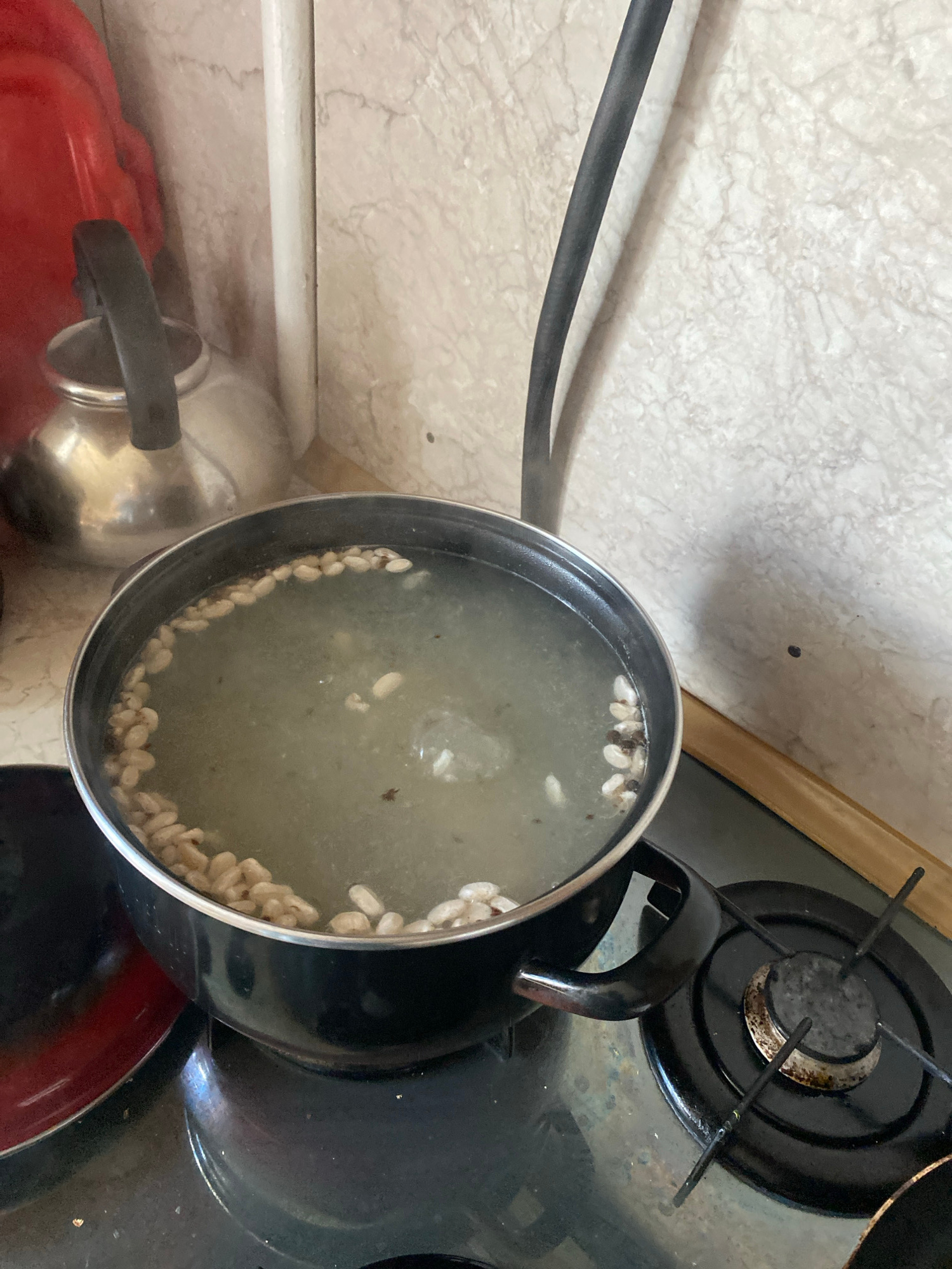 A close-up shot captures a pot of boiling water on a stovetop. The pot, black with a silver rim, contains a light-colored liquid with what appear to be chickpeas or white beans floating around the edges and throughout the water. A dark handle is visible on the right side of the pot. The stovetop is black with a circular burner, and the pot rests on the grates of the burner. To the left of the pot, a portion of a red object, possibly a container, is visible. In the background, a stainless steel kettle with a black handle sits on a counter. The wall behind the kettle has a marbled pattern. A thick black cable runs vertically down the right side of the frame against the marbled wall. The scene is indoors, likely in a kitchen, and the location is Krupki, Belarus.