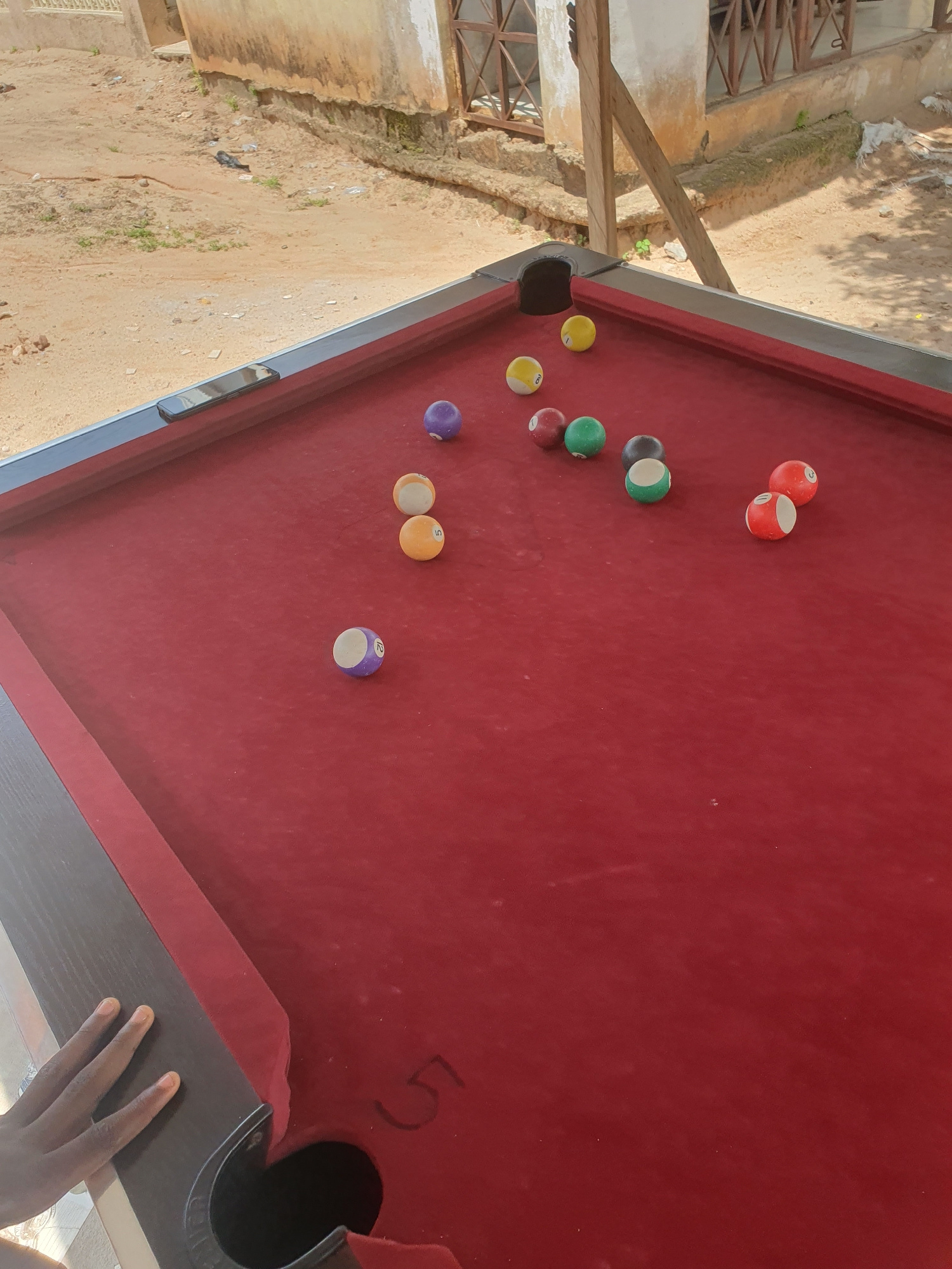 The image shows a close-up, high-angle view of a red pool table with several pool balls scattered across it. In the background, there is an outdoor setting with sandy ground, a building with weathered walls, and some wooden structures. There is a hand reaching into the frame from the bottom left, resting on the edge of the pool table. The scene is likely located in Lokoja, Nigeria, given the architectural style and the general environment depicted.