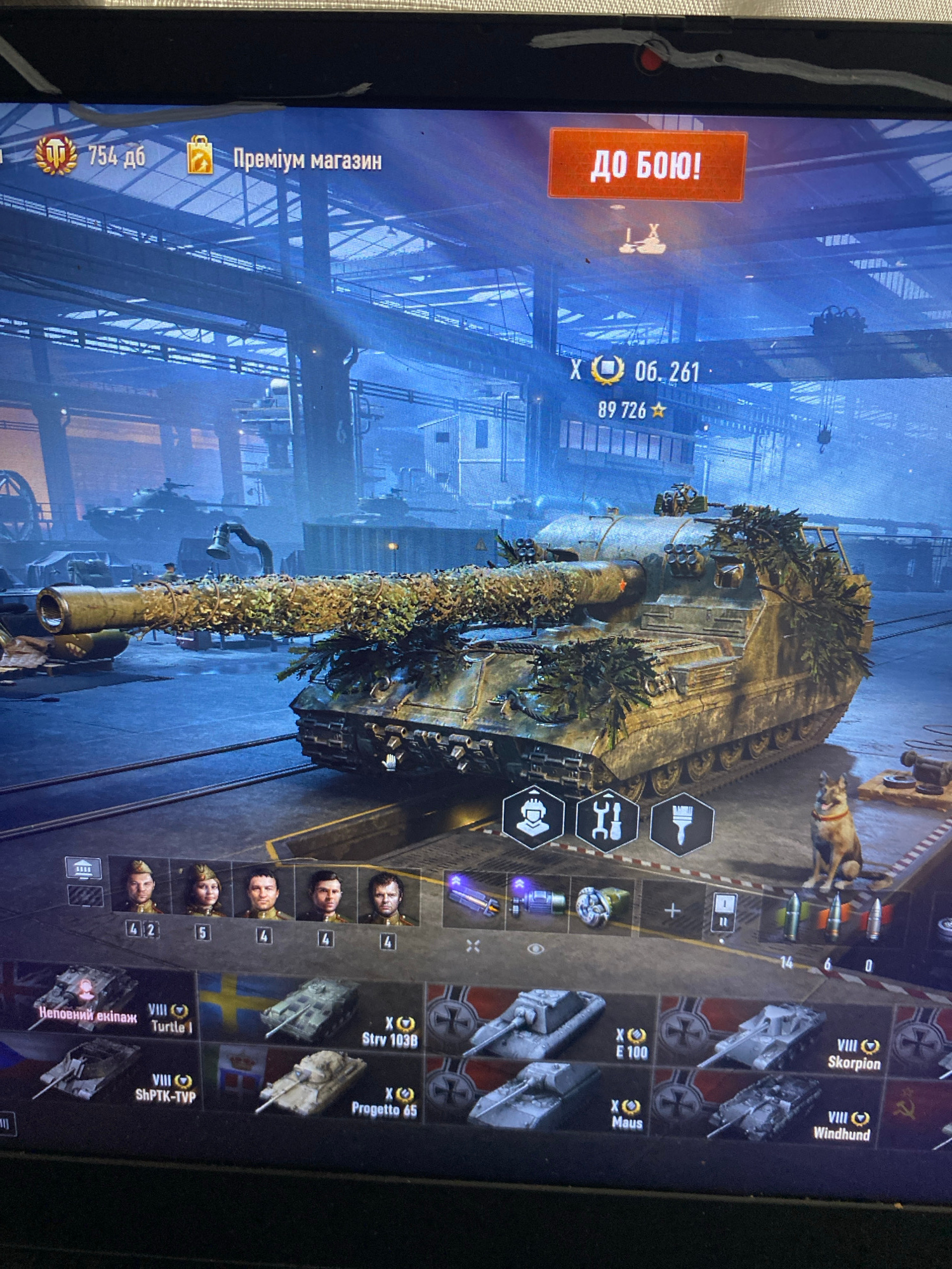 The image shows a screenshot of a video game interface, likely a tank simulation game. A heavily camouflaged tank is prominently displayed in the center. The interface includes icons for game actions, crew members, and a selection of other tanks. The top of the screen displays game currency and options like "Premium Shop" and "To Battle!" in Ukrainian. There is no visual information in the image that suggests a specific location in the city of Szczecin, Poland, or any real-world environment. The scene is entirely digital within the game's graphics.