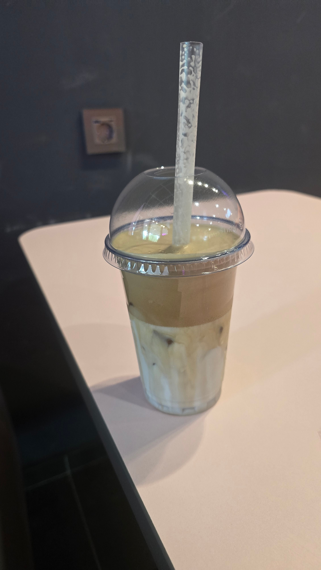 The image displays a clear plastic cup filled with a layered beverage, likely a coffee drink, topped with a domed plastic lid. A clear, textured plastic straw extends from the lid into the drink. The beverage itself exhibits distinct layers, with a lighter creamy color at the bottom, a darker brown layer in the middle, and a lighter, foamy layer at the top. Ice cubes are visible within the drink. The cup is positioned on a surface that appears to be a table or countertop, with a dark, reflective surface in the foreground and a lighter, possibly paper or fabric, surface behind the cup. The background on the left is a dark, textured wall with a small, framed picture. The lighting suggests an indoor setting, possibly a cafe or home. No specific time of day or weather conditions can be determined. The image is a close-up of the drink, with no people or specific activities explicitly shown. There is no visible text in the image. The provided location context of Ukhta, Russia, does not offer any direct visual cues within the image itself.
