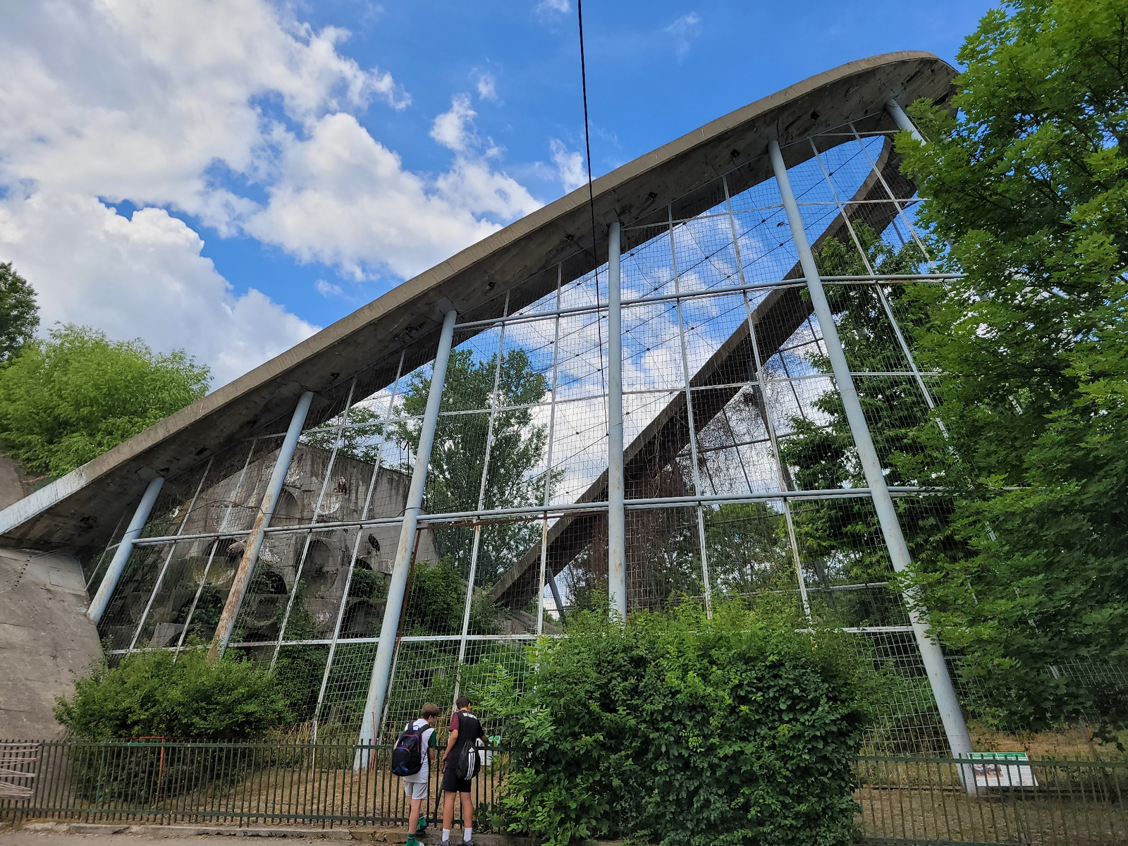 Two young people, one wearing a backpack, stand facing a large, angled structure covered with a mesh fence. The structure appears to be a building or enclosure with an open, concrete-like facade. Behind the mesh, there are hints of what looks like a rough, rocky interior with some vegetation growing. The sky above is blue with scattered white clouds, and lush green trees flank the right side of the structure. A dark, thin cable hangs vertically in the middle of the frame. The ground in front of the enclosure is visible, with a metal fence separating the observers from the area. The overall impression is that of an outdoor exhibit or zoo enclosure.