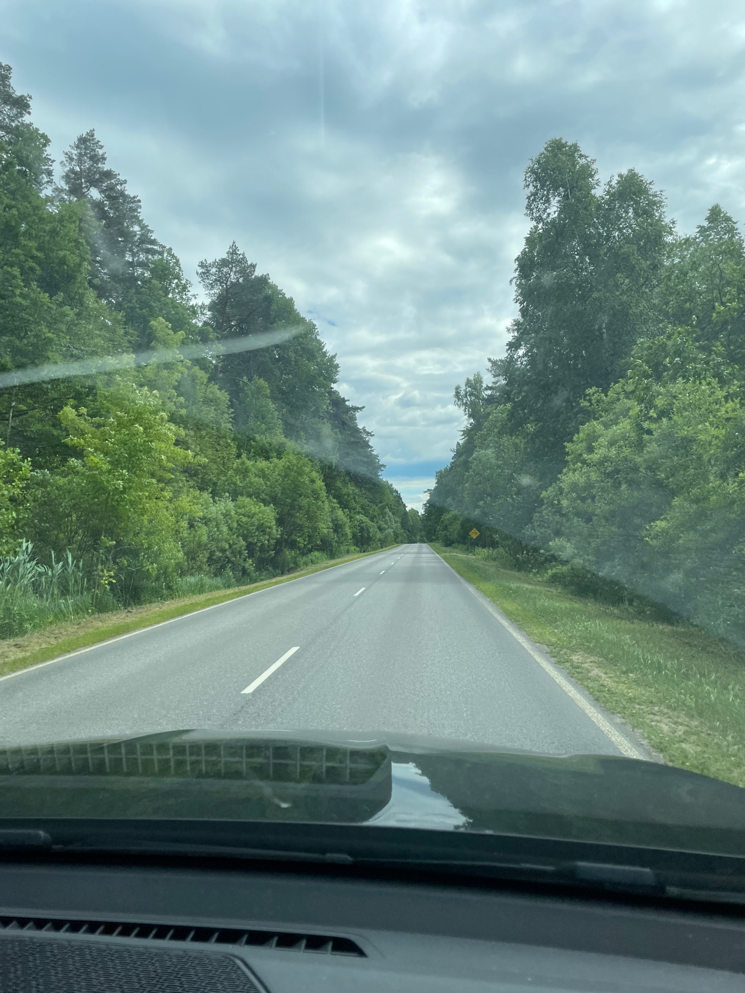 An eye-level, forward-facing, full shot captures a paved road stretching into the distance, bordered by lush green trees on both sides. The sky above is overcast with a mix of gray and white clouds, suggesting a temperate climate. The road is marked with white lines, indicating a two-lane highway. In the foreground, the reflection of the sky and trees is visible on the dashboard of a vehicle, indicating the photo was taken from inside a car. There is a yellow road sign visible on the right side of the road in the distance, but its details are unclear. The environment appears to be rural, with dense vegetation lining the road.