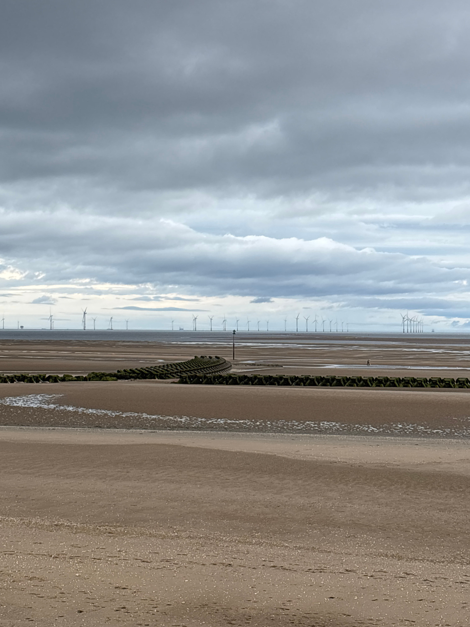An eye-level, wide shot captures a vast, grey, cloudy sky dominating the upper half of the frame. Below, the horizon showcases an offshore wind farm, with numerous white turbines dotting the calm sea. The foreground is a wide, sandy beach, marked by the receding tide. A line of dark, textured breakwaters runs across the mid-ground, curving slightly to the right. A single person is visible in the distance, wading in shallow water on the beach.