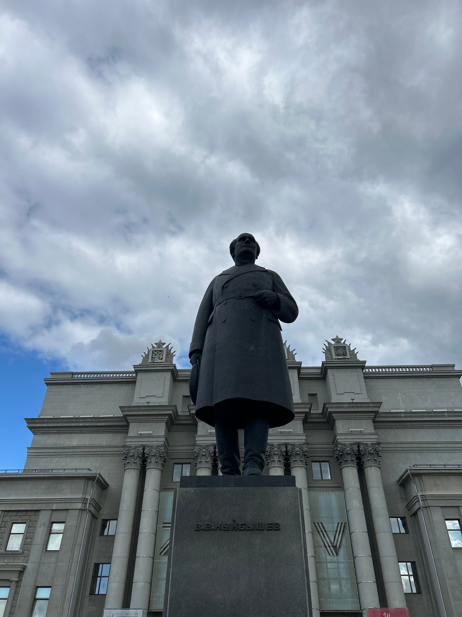 A low-angle, medium shot captures a dark, imposing statue of a man in a long coat, standing on a stone pedestal. The statue is centrally positioned against a backdrop of a large, grey, neoclassical-style building with imposing columns and decorative elements on its roof. The sky above is a dramatic mix of heavy grey clouds with patches of bright blue peeking through. The inscription on the pedestal, rendered in dark, raised lettering, appears to read "В.В. КУЙБЫШЕВ," identifying the figure as V.V. Kuybyshev. This monument is located in Novokuybyshevsk, Russia, in front of the Kuybyshev Azot industrial complex. The scene is static and solemn, with no people or animals visible other than the sculpted figure.