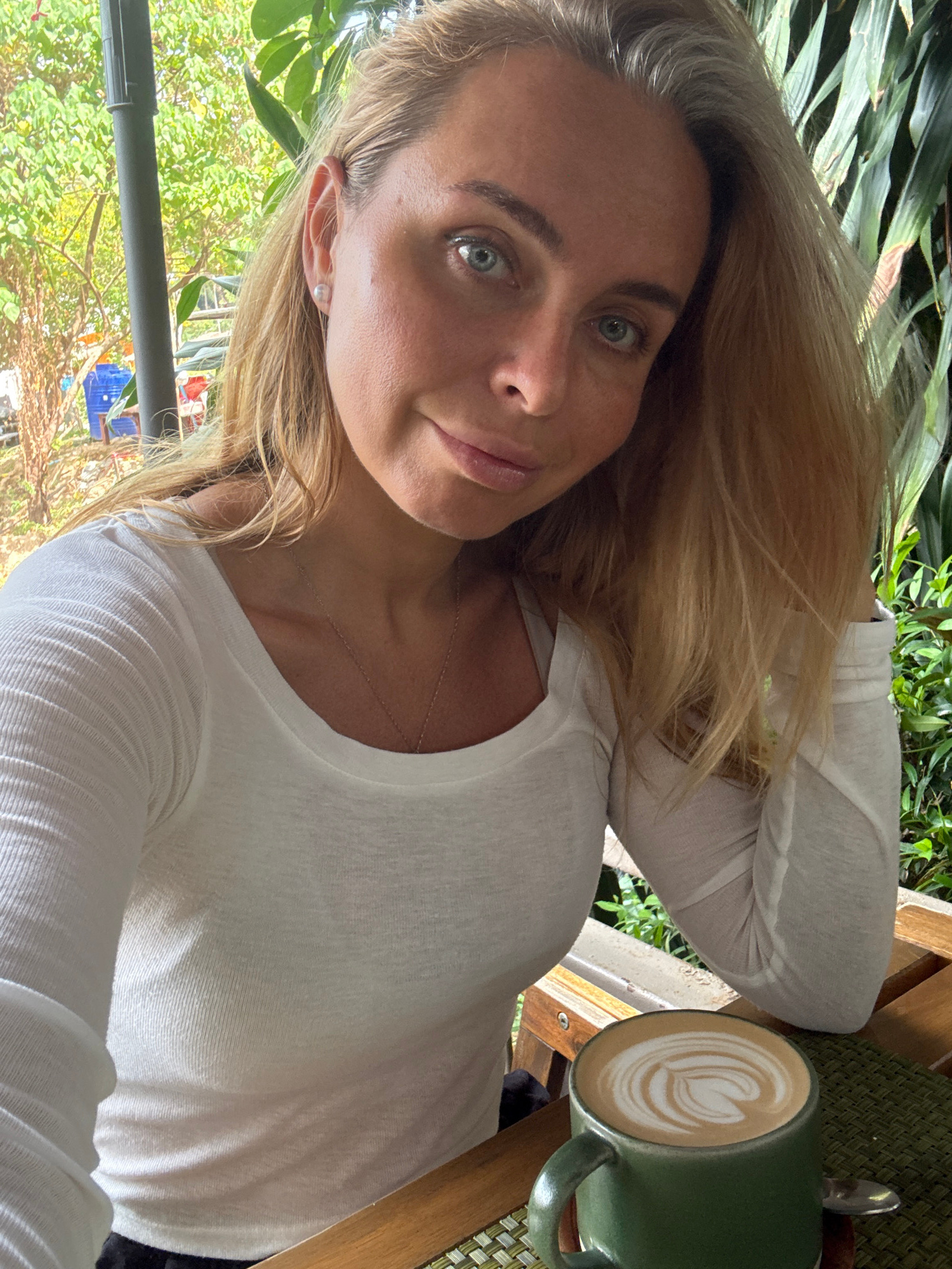 A medium shot photograph, taken from a slightly low angle, captures a fair-skinned woman with blonde hair and blue eyes posing for a selfie at an outdoor cafe in Baan Chaweng Noi, Thailand. She is wearing a white long-sleeved ribbed top and a delicate necklace. Her left elbow rests on a wooden table, and a green mug filled with a latte sits in front of her. The background features lush green foliage and a glimpse of what appears to be part of a blue plastic container. The lighting suggests it is daytime, with a warm, natural glow illuminating the scene.