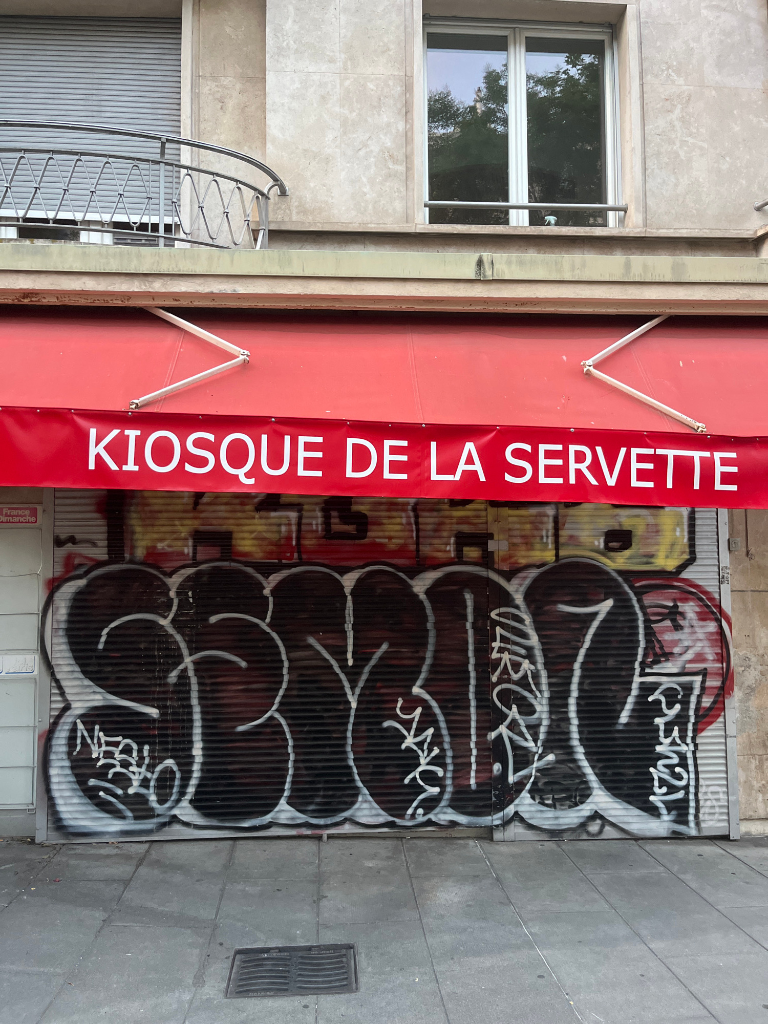 This image shows a closed storefront with a red awning. The awning has white lettering that reads "KIOSQUE DE LA SERVETTE". The storefront itself is a metal rolling door covered in graffiti. The graffiti is in various colors, including black, white, yellow, and red, and appears to be spray-painted tags and bubble letters. Above the awning, there is a building with a balcony and a window. The building's facade is made of stone. The ground in front of the storefront is paved with gray tiles. A metal grate is visible in the pavement. The scene appears to be an urban environment. There are no people visible in the image.