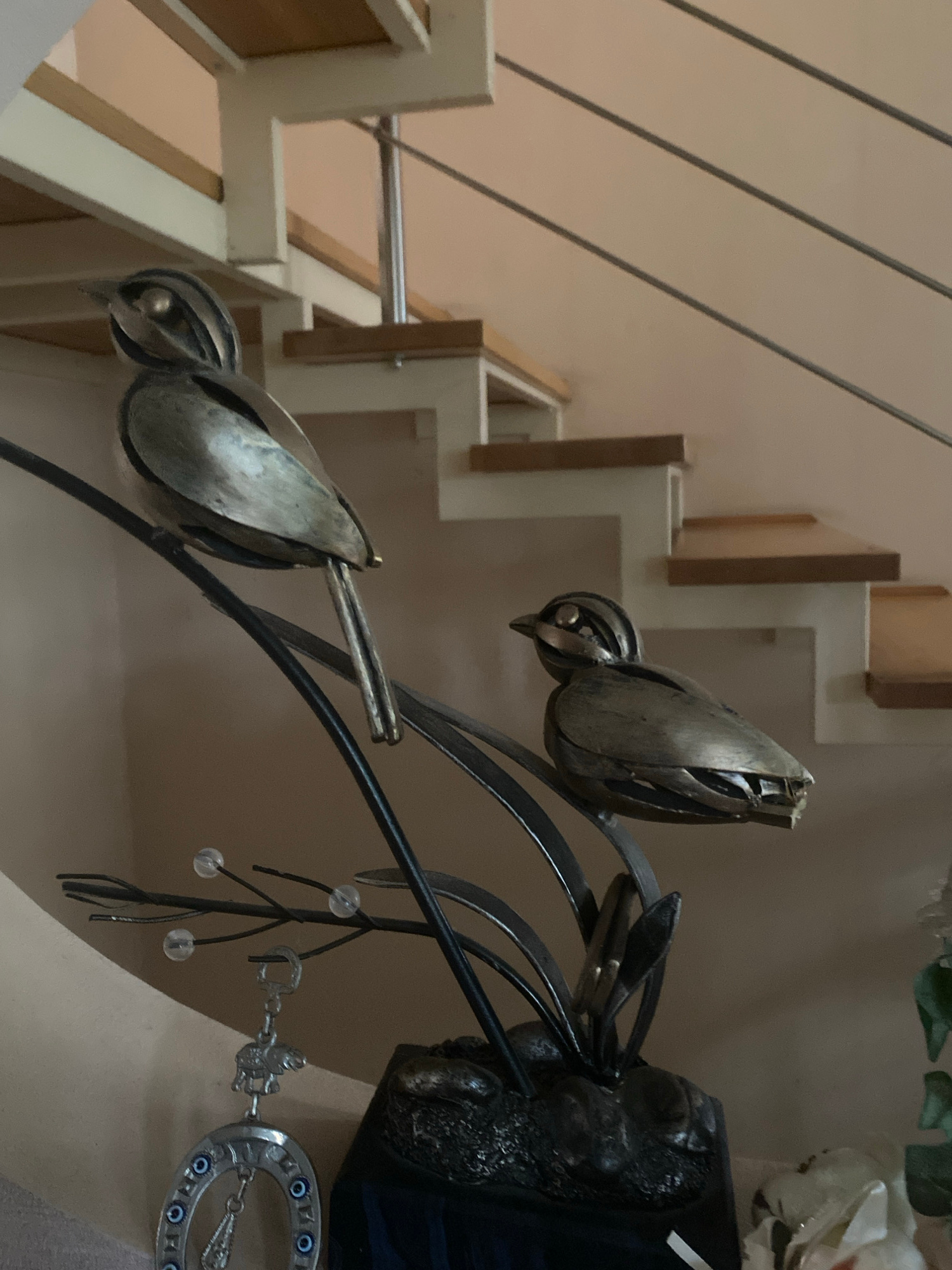 The image shows a decorative metal sculpture of two birds perched on a branch. The sculpture is placed in an indoor setting, likely a home, with a staircase visible in the background. The birds are metallic and appear to be made of brushed or polished metal, with detailed feathering and eye features. One bird is larger and positioned on the left, while a smaller bird is on the right. The branch is also made of dark metal and has small, clear, bead-like embellishments that could represent dew drops or berries. A hanging charm with a horseshoe and an elephant figurine is attached to the branch. The overall ambiance suggests a decorative display in a residential interior. There is no indication of the city of Kharkiv, Ukraine, in the image.
