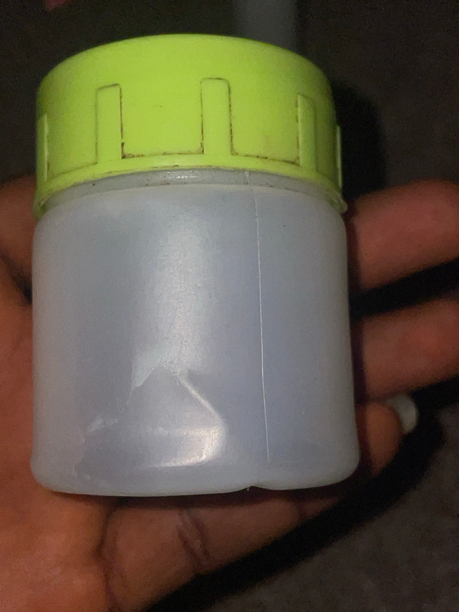 A close-up, slightly blurry image shows a translucent white plastic bottle with a bright lime-green ridged cap held in a dark-skinned hand. The bottle appears to be filled with a clear liquid. The background is dark and indistinct, suggesting an indoor setting or nighttime. The image is a product shot, possibly for identification or sale. The exact location in Jalingo, Nigeria is not discernible from the visual information provided.