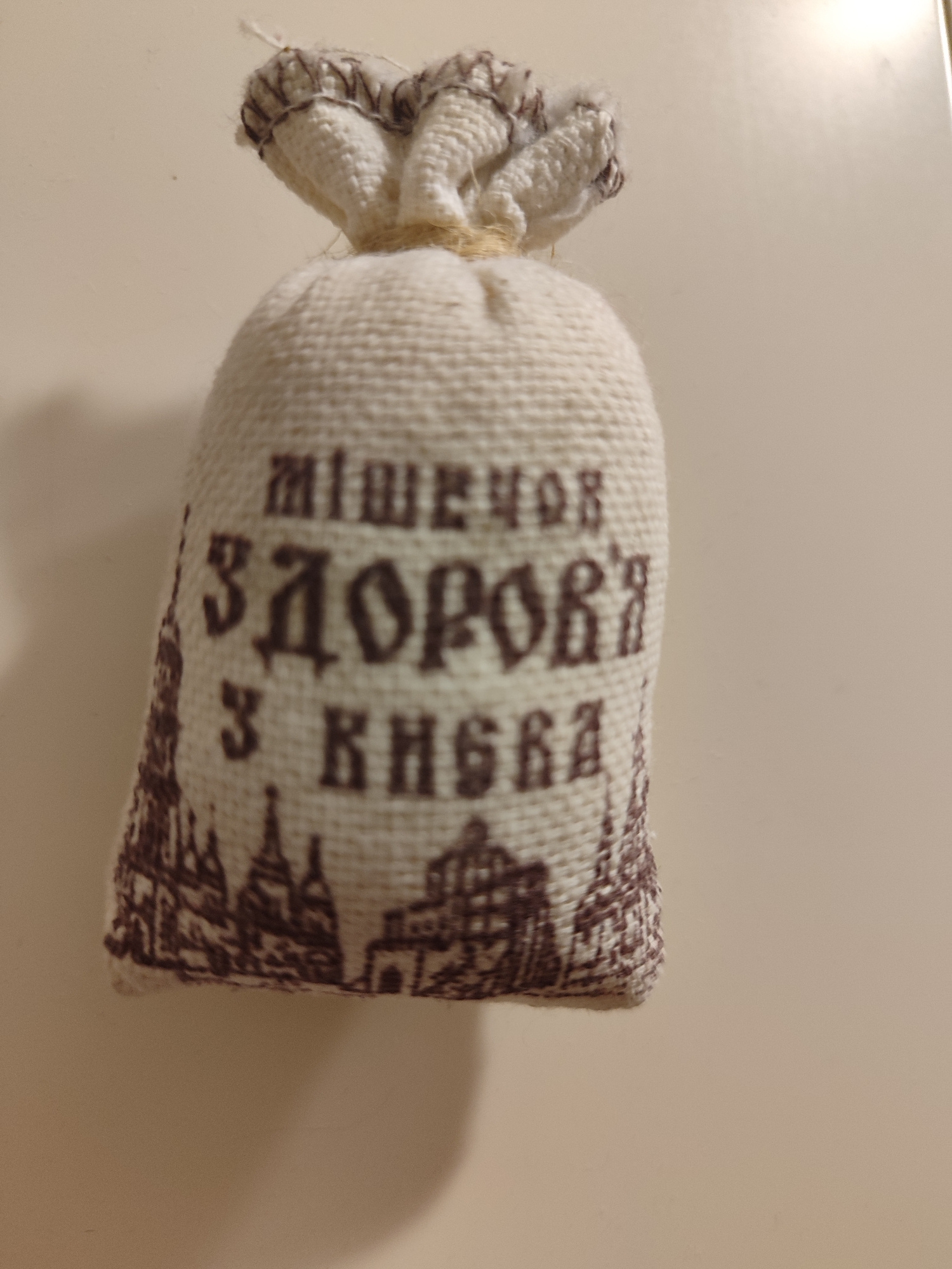A small, burlap pouch with a drawstring closure sits upright on a light-colored, smooth surface. The pouch is white with a textured weave and features brown lettering and an illustration. The lettering appears to be in Cyrillic script, and the illustration depicts a skyline with several buildings and spires. The pouch is illuminated from the top right, casting a soft shadow to the bottom left. The background is a plain, neutral color. There is no indication of the city of Mykolaiv, Ukraine within the image itself.