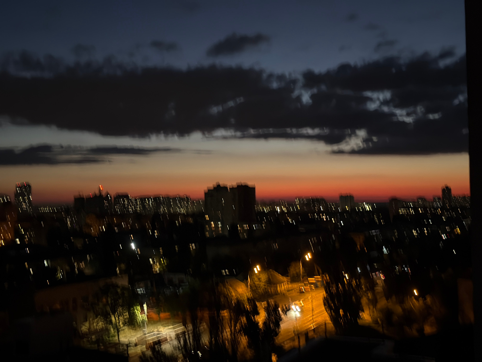 The image captures a blurry twilight cityscape, likely from an elevated viewpoint in Kyiv, Ukraine. The sky transitions from a deep blue at the top to a warm orange and red hue near the horizon, indicating sunset or sunrise. Dark, elongated clouds stretch across the upper portion of the sky. Below the sky, the silhouettes of numerous buildings, ranging in height, are visible, their windows illuminated by artificial lights, creating a speckled effect. Streetlights cast pools of yellow light on what appear to be roads and pathways in the foreground, with the blurred streaks of moving vehicles suggesting traffic. Trees with dark foliage are interspersed throughout the urban landscape. The overall impression is one of a city transitioning from day to night, with the blurriness suggesting a long exposure or motion blur.