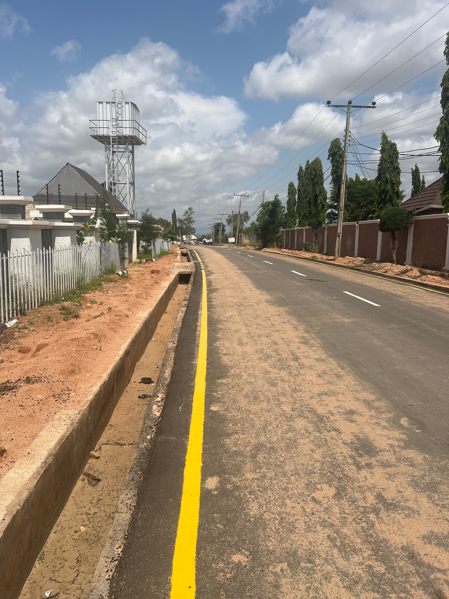 The image captures a view of a street in Howayi, Nigeria, under a partly cloudy sky. The street is paved and features a bright yellow line marking the edge of the road. To the left of the road, a concrete drainage ditch runs alongside a fence, which appears to be made of white vertical bars. Beyond the fence, a building with a gray, pitched roof is visible, and to its left, a tall, silver water tower stands prominently. The right side of the road is bordered by a wall, a row of tall, slender trees, and more buildings with brown, tiled roofs. In the distance, several utility poles with electrical wires are also visible. The overall scene depicts a residential or developing urban area.