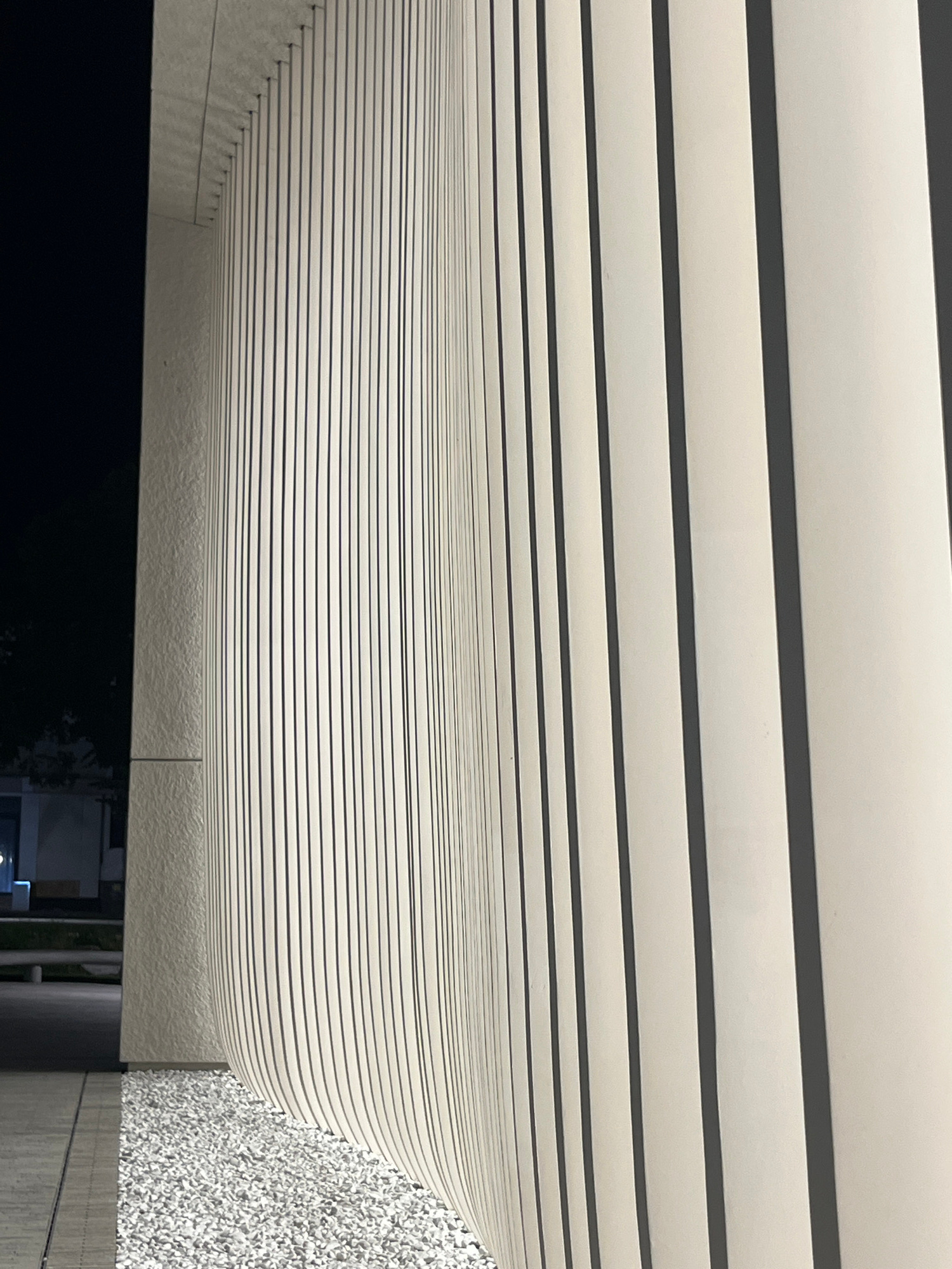 The image depicts a modern architectural facade with vertical elements, likely louvers or blinds, rendered in a pale, creamy color with dark gray or black shadowing in between. These elements are arranged in a repetitive pattern, creating a sense of depth and rhythm. The facade appears to be part of a building situated in an outdoor urban environment at night. To the left of the facade, a dark, shadowed area suggests the night sky and possibly some distant trees. Below the facade, a section of light-colored gravel is visible, and further to the left, a paved walkway and what appears to be a low wall or bench are discernible. The overall impression is one of a contemporary design element in a nighttime urban setting. The image does not contain any people or identifiable actions. The location is specified as Almaty, Kazakhstan, suggesting this architectural feature is part of a building within that city.