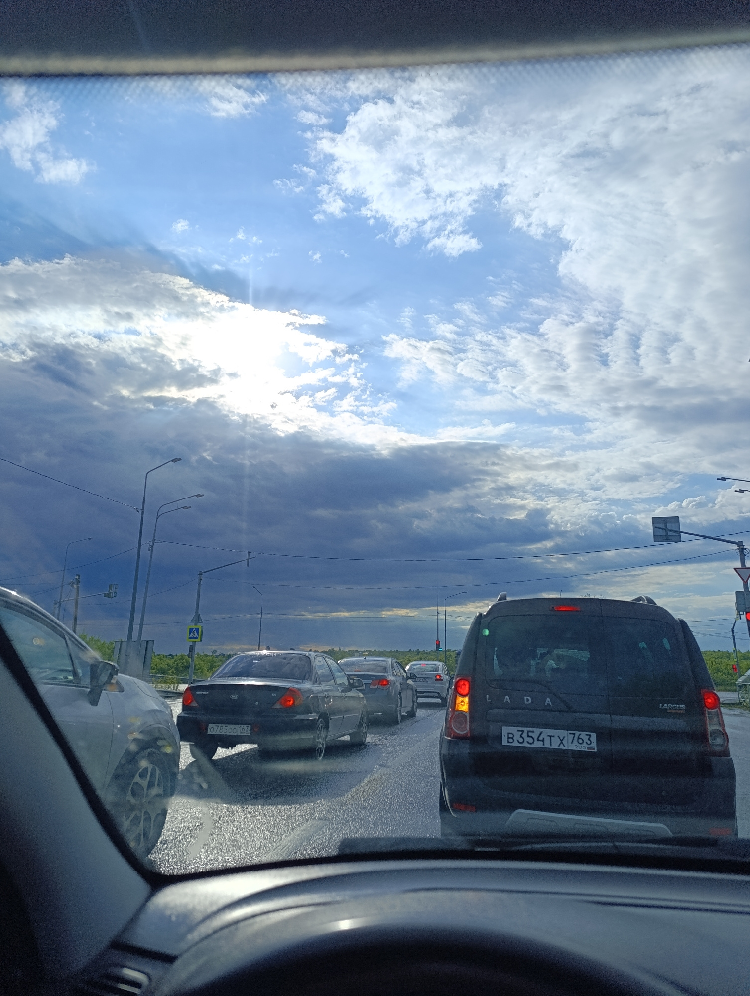 The image shows a view from inside a vehicle, looking out through the windshield. The vehicle is in traffic on a wet road, indicating recent rain or current precipitation. Ahead, several other cars are visible, all stopped or moving slowly. The sky is overcast with dark clouds, but patches of bright blue sky and sunlight break through, casting rays of light. Streetlights and traffic signals are present along the roadside. The overall scene depicts a traffic jam in a semi-urban or suburban environment, with a focus on the road, vehicles, and the sky.