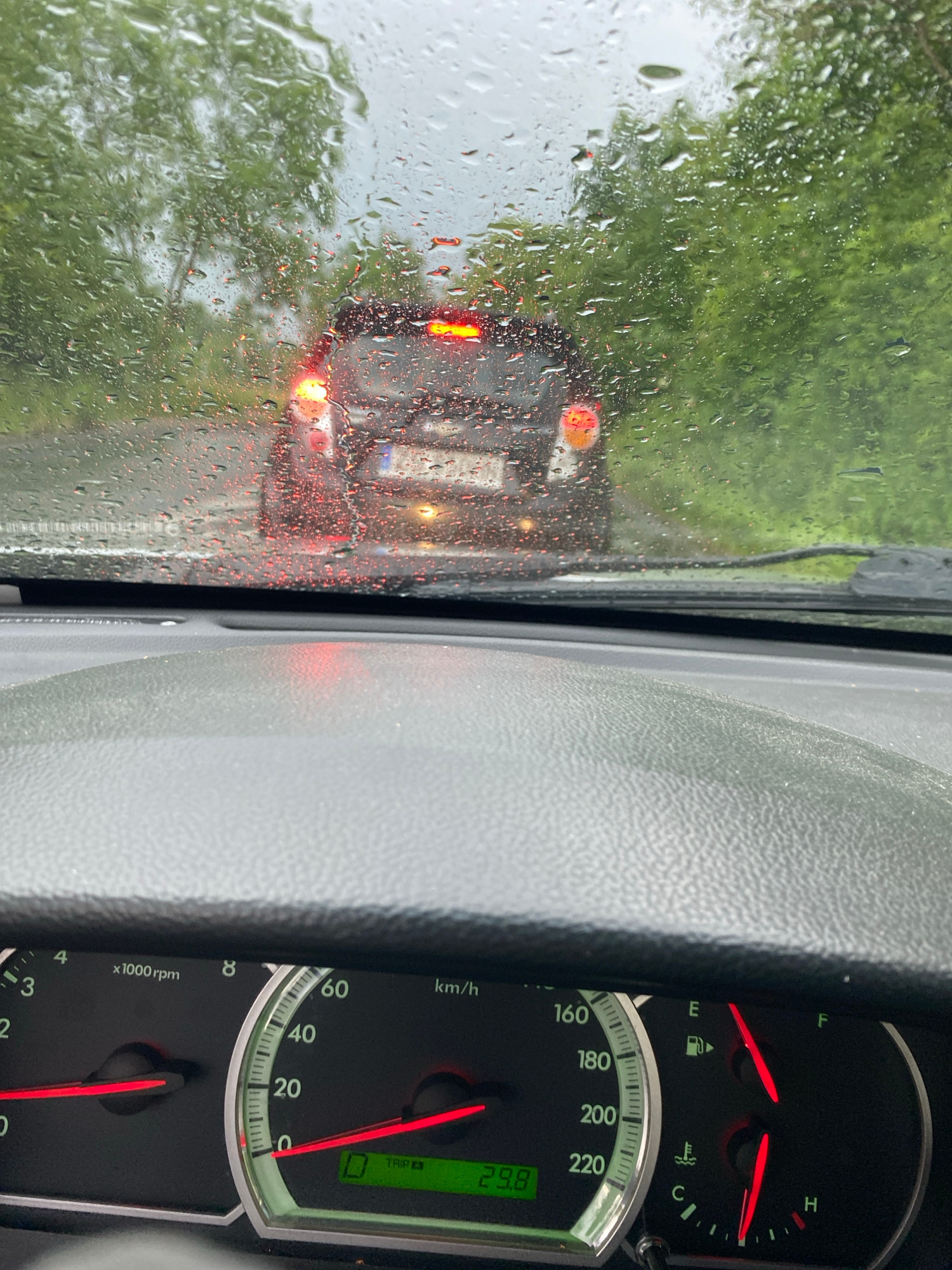 The image shows a view from inside a car during heavy rainfall, looking forward through the wet windshield. Raindrops are visible on the glass, distorting the view of the road ahead where traffic is backed up. The red taillights of the car in front are illuminated, indicating it's likely a cloudy or dark day, exacerbated by the rain. Green foliage from trees lining the road can be seen through the rain-streaked window.
Inside the car, the dashboard is visible on the right side of the frame. It includes various gauges, such as a speedometer with markings up to 220 km/h, a tachometer marked in x1000 rpm, a fuel gauge, and a temperature gauge. A digital display shows "29.8". The textured interior of the car is also visible. The setting is a road, likely in or around Szczecin, Poland, as suggested by the context. The overall mood conveyed is one of a damp, possibly slow commute or journey due to the weather conditions. The time of day is not explicitly indicated but the overcast sky and illuminated taillights suggest it's not bright daylight.
The visible text includes: "2", "3", "4", "x1000 rpm", "8", "60", "km/h", "E", "F", "40", "160", "180", "20", "200", "220", "TRIP", "29.8", "C", and "H".