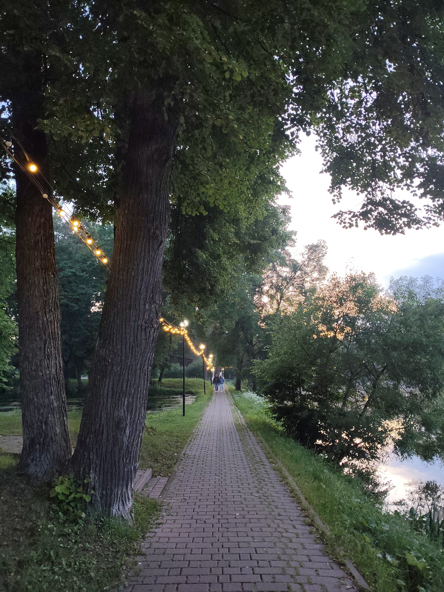 The image depicts a paved pathway lined with trees and illuminated by a string of warm-toned lights. The pathway runs alongside a body of water, likely a river or lake, with lush green vegetation on the opposite bank. In the distance, two figures can be seen walking along the path. The overall atmosphere is serene and natural, with the lighting suggesting it is dusk or early evening. The location is identified as being in the city of Chekhov, Russia.