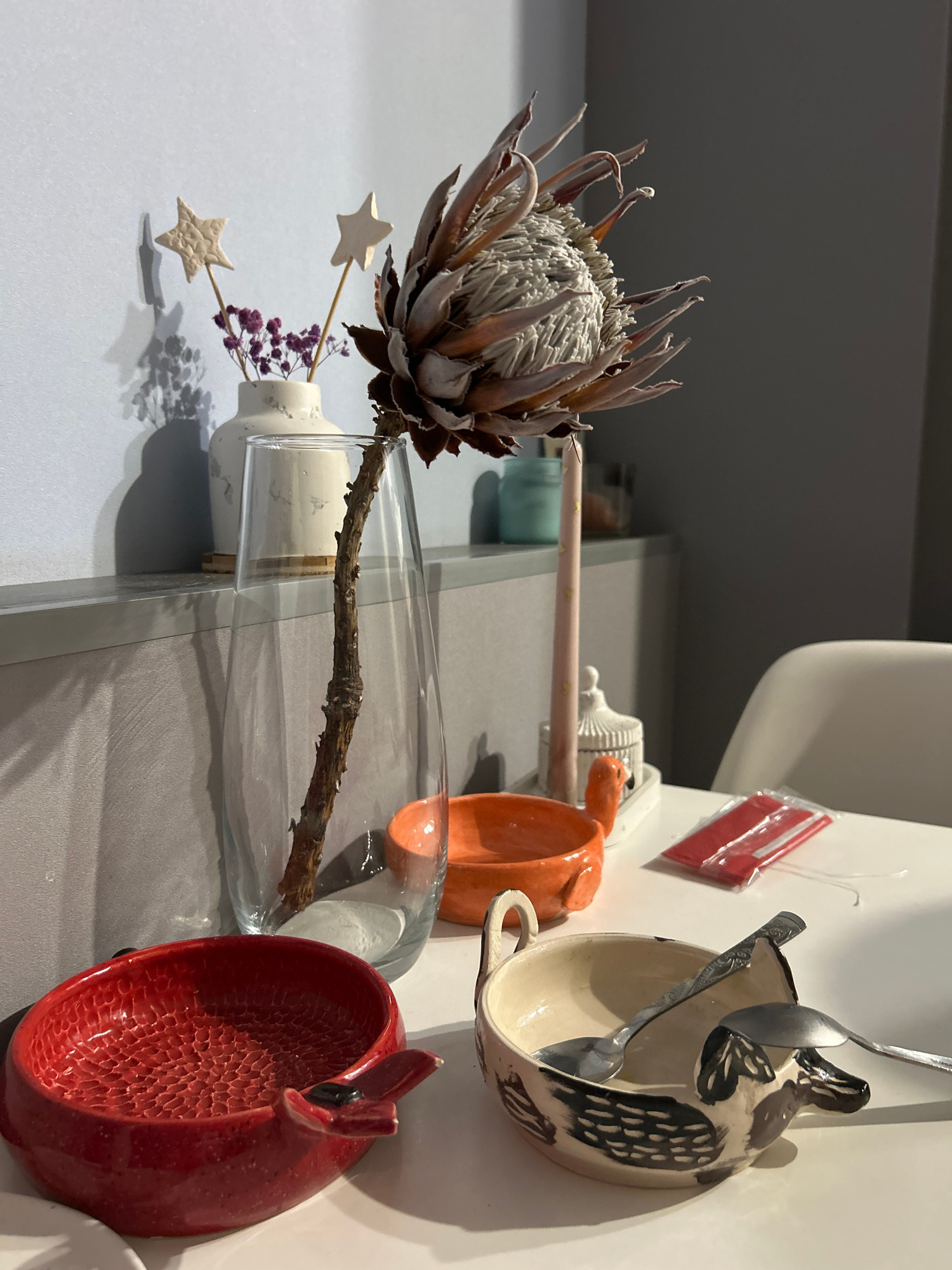The image shows a still life composition on a white surface, likely a table, with a muted grey and blue background. In the foreground, a red ceramic bowl with a textured interior and a whimsical fin-like handle sits to the left. To its right is a cream-colored ceramic bowl decorated with a black and brown dog-like figure and two spoons resting inside. Behind these bowls, an orange ceramic bowl with handles is visible. A tall, clear glass vase contains a dried protea flower with a long, woody stem. To the left of the vase, a white ceramic vase holds a few sprigs of dried purple flowers and two small star-shaped decorations on sticks. A tall, pink candle with gold star accents stands near a small, white decorative birdhouse. In the background, a glimpse of a white chair and a red package are visible. The scene is lit by natural light, casting shadows. There are no people present in the image. The location is identified as Orenburg, Russia.