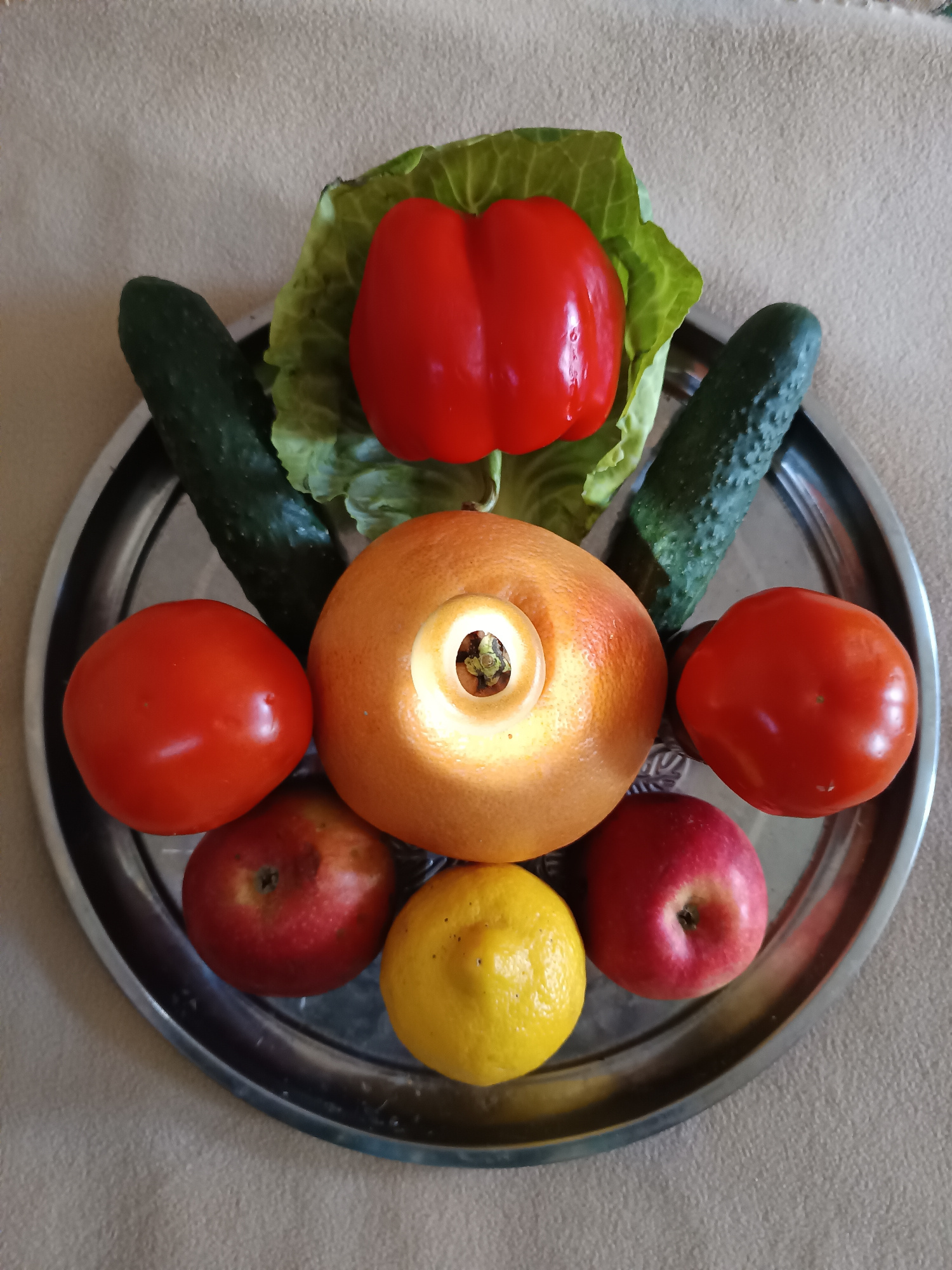 A high-angle, medium shot captures a metallic tray filled with assorted fruits and vegetables arranged on a textured, light-brown surface. The arrangement is symmetrical, with two cucumbers positioned vertically on either side of a red bell pepper resting on a bed of lettuce leaves. In the center, a grapefruit is positioned with a light source emitting from its hollowed-out center. Flanking the grapefruit are two red tomatoes. Below the grapefruit, a yellow lemon is centered between two red apples. The metallic tray has a rim with decorative etching. The lighting appears to be coming from overhead, casting shadows on the fruits and vegetables. The scene is static, with no discernible actions or interactions taking place.