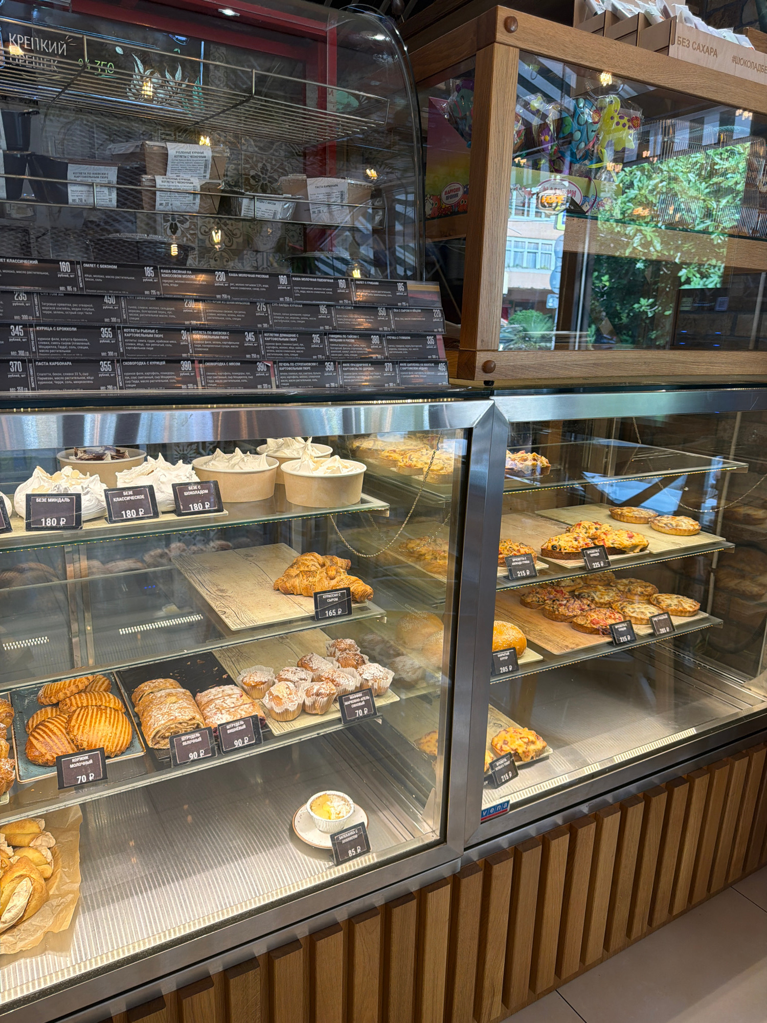 This image depicts a bakery display case filled with various pastries, breads, and sweets. The display is illuminated and made of glass, allowing a clear view of the products. In the background, there are shelves stocked with packaged goods. The overall environment appears to be a retail establishment. The price tags are in Russian rubles, and the signage suggests the location is Sochi, Russia. Some products are labeled with Russian text, including descriptions and prices. For example, one label reads "КРЕПКИЙ" and "350" indicating a strong product priced at 350 rubles. Another label says "БЕЗ САХАРА" (sugar-free). The display features items like croissants, tarts, muffins, and layered pastries. The shelves are organized with some products in individual containers and others displayed loosely. The lighting highlights the textures and colors of the baked goods.