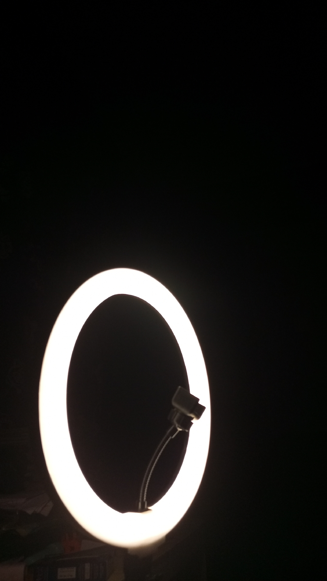 A bright, circular light illuminates a dark background. A black, flexible arm extends from the bottom of the circle, holding a small, white object at its end. The lower portion of the image shows a dark, cluttered area with some indistinct objects and shapes, suggesting it is an indoor space.