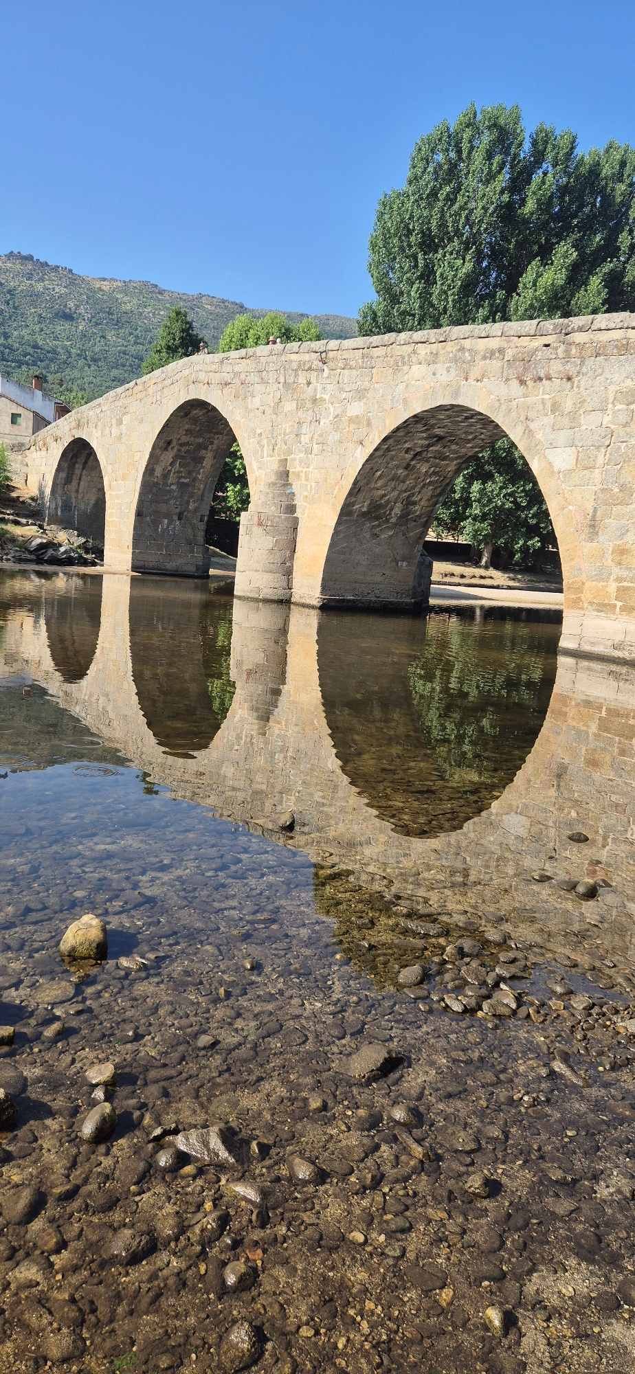An eye-level, vertical shot captures a stone bridge spanning a shallow river under a clear blue sky. The bridge, constructed from rough-hewn, light brown stones, features three large, rounded arches. Sunlight illuminates the bridge, casting distinct reflections on the calm water's surface. The riverbed is visible through the clear water, revealing a multitude of stones and pebbles of varying sizes and colors, with some larger rocks dotting the scene. Lush green trees flank the bridge on both sides, with a hillside covered in vegetation and trees visible in the background. A building with a white wall and a reddish-brown roof is partially visible to the left of the bridge. Several people, too small to discern details, are visible on top of the bridge.
