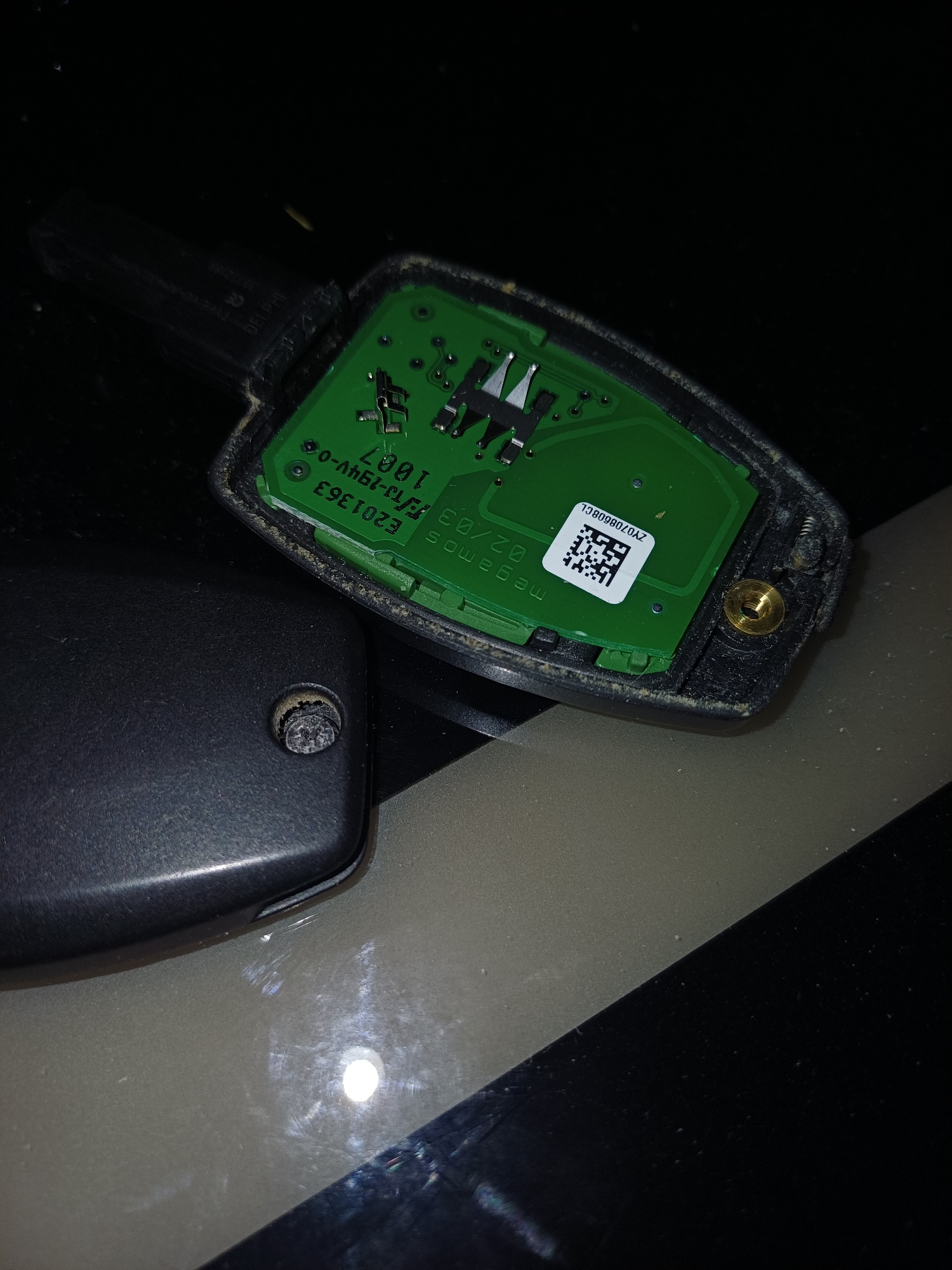 The image shows the disassembled electronic components of a car key fob. The green printed circuit board (PCB) is visible, with various electronic components soldered onto it. A black plastic casing for the key fob is also present, with a screw hole and a small screw visible. There is a small white sticker with a QR code and text. The overall scene is dimly lit, with the objects resting on a surface that appears to be a table or desk. The image does not contain any information or context that would allow for the determination of a specific location in Middlesbrough, United Kingdom.
