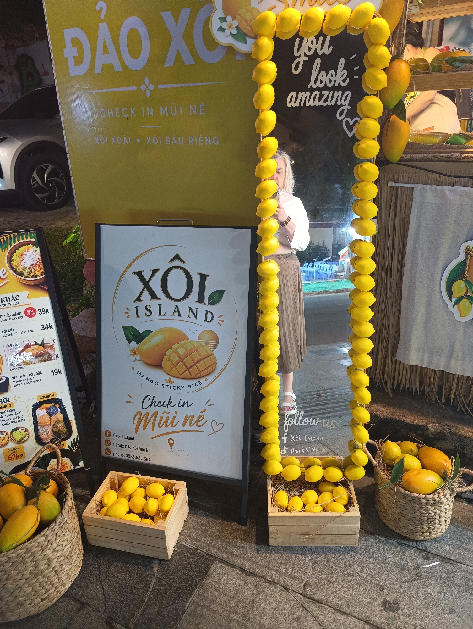 The image depicts an outdoor scene, likely a food stall or shop, with a focus on mango sticky rice. A large yellow banner with Vietnamese text "ĐẢO XÔI" and "CHECK IN MŨI NÉ" dominates the background. To the left, a menu board advertises various sticky rice dishes, including "XÔI XOÀI" (Mango Sticky Rice) and "XÔI SẦU RIÊNG" (Durian Sticky Rice). A woman in a white top and a beige skirt is standing near a reflective surface adorned with a string of yellow, lemon-like decorations and the text "you look amazing". In the foreground, two woven baskets and a wooden crate are filled with yellow fruit, resembling lemons or small mangoes, arranged on a stone-paved ground. The overall impression is a vibrant and inviting setup promoting a local delicacy. The location is identified as Phu Thuy Ward, Vietnam.