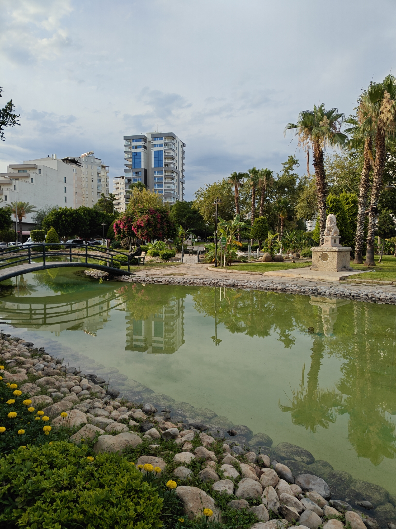 This image captures a serene park scene in Antalya, Turkey, featuring a reflective pond with a small arched bridge. The pond's green water mirrors the surrounding environment, including tall palm trees, lush green foliage, and modern buildings in the background. On the right side of the pond, a stone pedestal holds a lion statue, adding an artistic touch to the landscape. Yellow flowers and dense green bushes line the pond's rocky edge in the foreground, contrasting with the smooth, paved walkway and grass further back. The sky above is partly cloudy, suggesting it could be late afternoon or early evening. The presence of apartment buildings suggests the park is situated within a developed urban area of Antalya. There are no people visible in the frame.