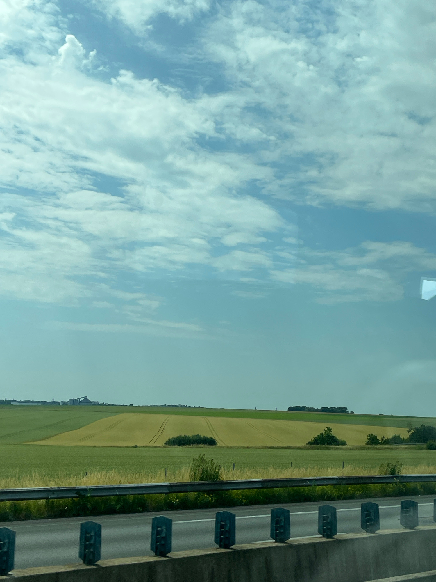 The image depicts a rural landscape with rolling fields under a cloudy sky. In the foreground, a concrete barrier and a metal guardrail line a road. Beyond the road, fields of varying shades of green and yellow stretch towards a distant horizon. A few trees are scattered across the landscape, and faint structures are visible in the far distance. The overall impression is one of an agricultural area. The specific location in the city of Eswars, France, is not discernible from the visual information alone.