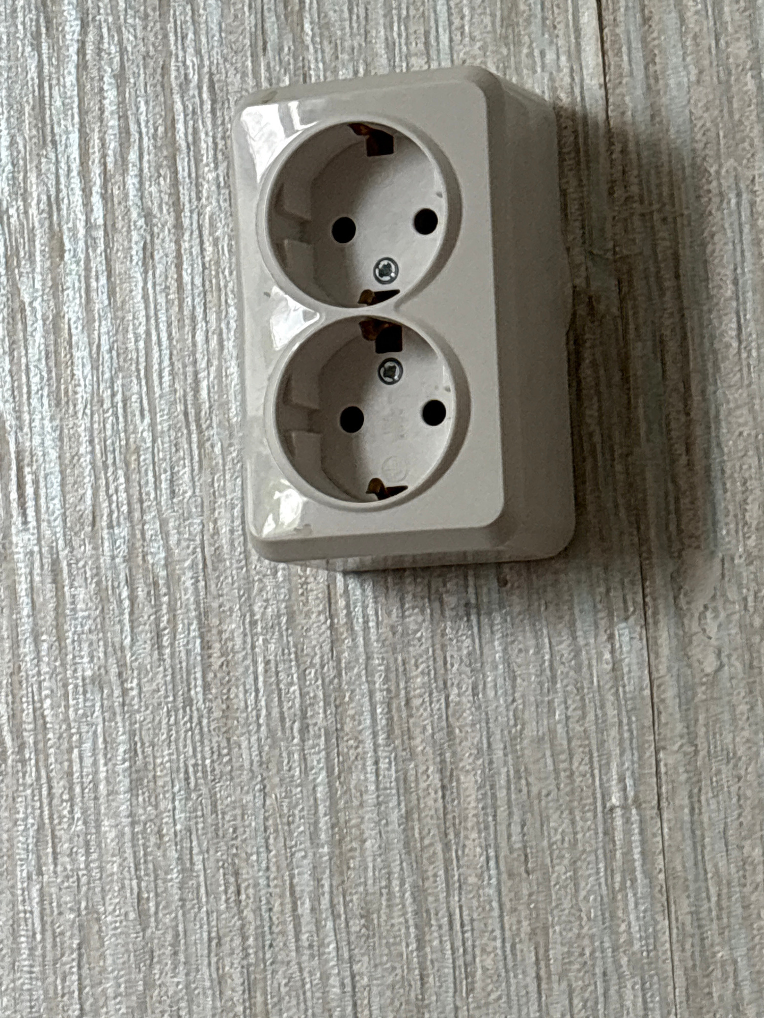 An off-white double electrical outlet is mounted on a textured light-colored wall. The outlet has two circular receptacles, each with a central hole and two slots for prongs. The wall surface has a vertical, ribbed texture. The scene is indoors and the lighting is neutral, indicating it is likely daytime or a well-lit interior space. There are no people or other objects visible in the frame.