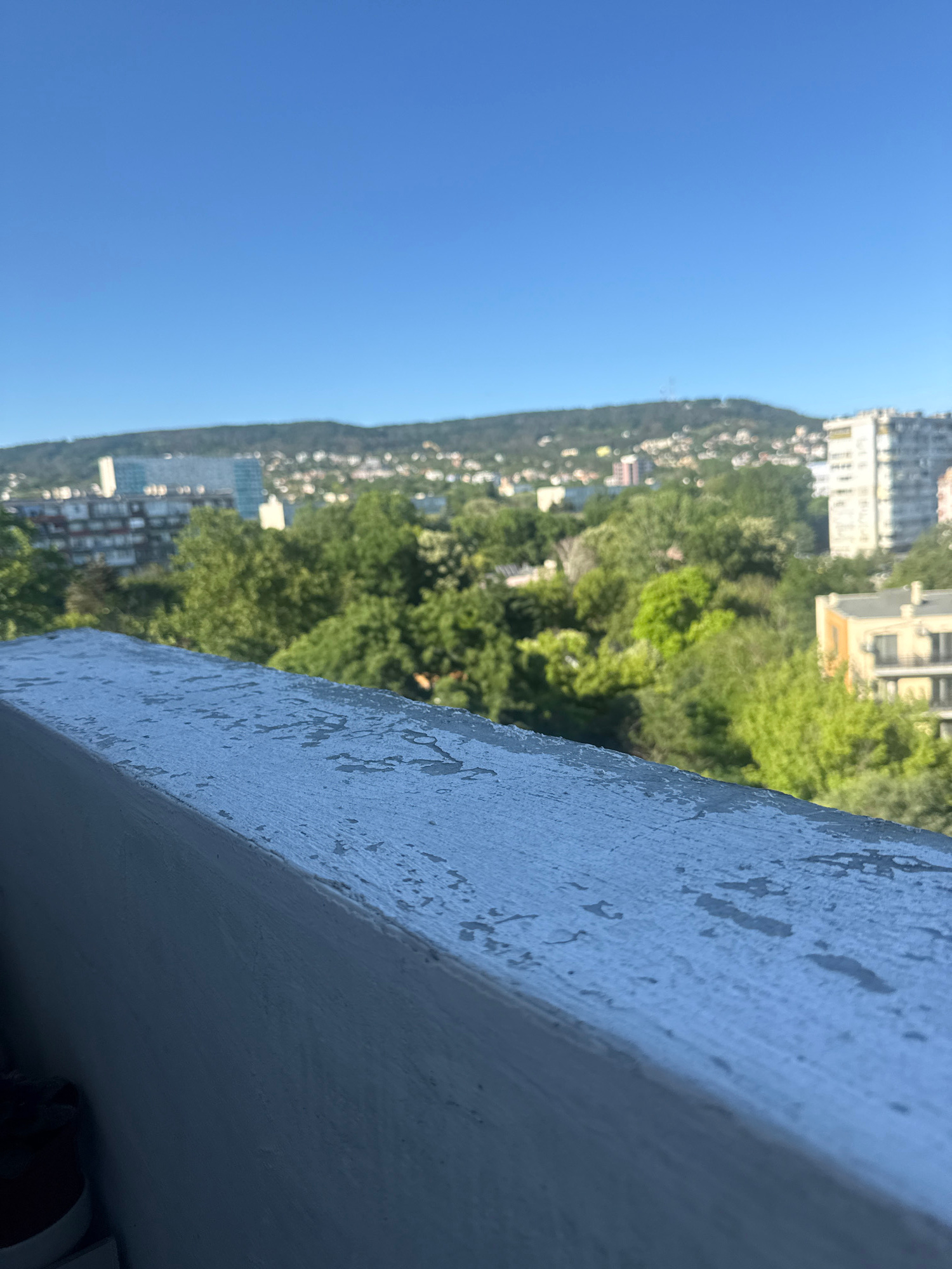 This image captures a view from a balcony or elevated outdoor space, looking out over a city with a hilly backdrop. In the foreground, a weathered, light-colored railing with peeling paint runs diagonally across the frame, its texture prominently featured. Beyond the railing, lush green trees form a dense canopy, interspersed with buildings of varying heights and architectural styles. Some buildings appear to be residential blocks with multiple floors, while others are taller, modern structures. The background is dominated by a verdant, sloping hill covered in scattered buildings, suggesting a town or city built on the landscape. The sky is a clear, bright blue. The scene suggests a daytime view, likely in an urban area that incorporates significant natural elements. Given the context provided, this scene is located in Varna, Bulgaria, a coastal city known for its hills and greenery overlooking the Black Sea.