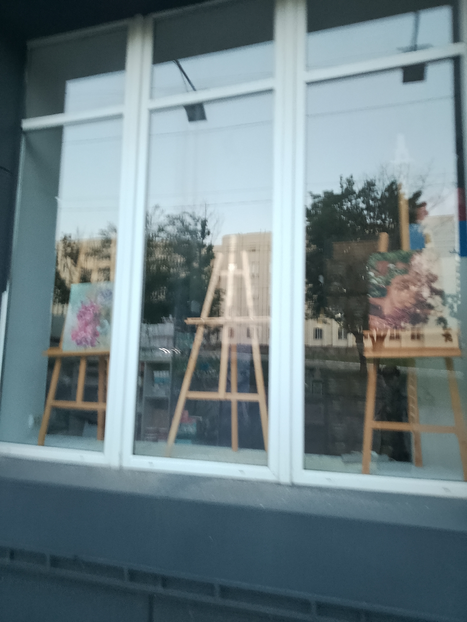 The image shows a storefront window with a view into an art display. Two wooden easels are visible, each holding a painting. The painting on the left features pink and white flowers against a light background. The painting on the right is partially obscured by reflections and appears to be a floral still life with shades of pink, purple, and green. In the background, trees and buildings are visible, suggesting an outdoor environment reflected in the window. The overall lighting indicates it is daytime. There are no people clearly visible. The building itself has a dark grey facade below the window, and a brick base. The window is divided into three panes by white mullions. This scene is located at a shop on Karl Maks (now Korolenka) Street in Dnipro, Ukraine.