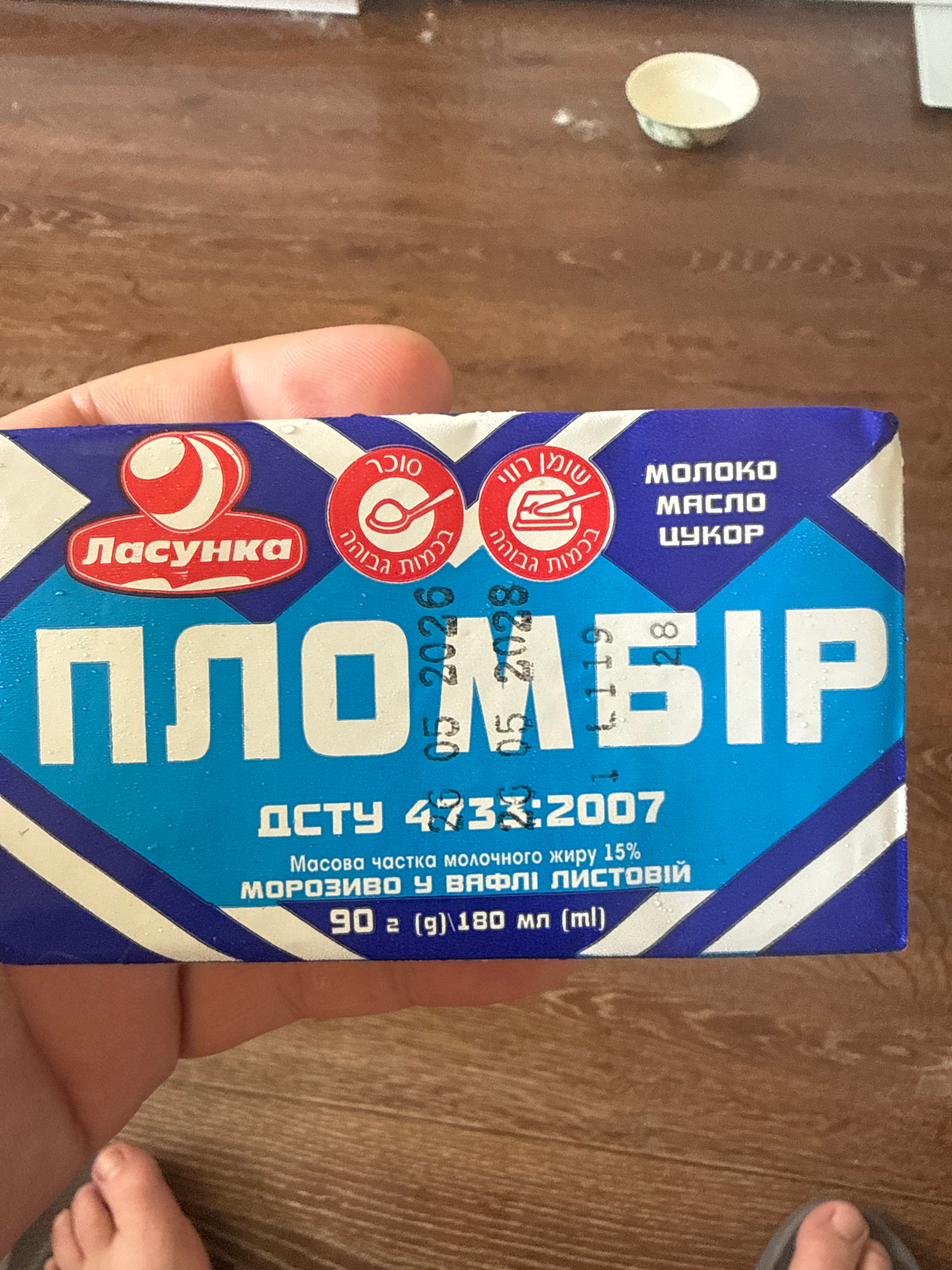 A hand holds a rectangular blue and white packaged product, possibly ice cream or butter, with prominent text in what appears to be Cyrillic and possibly Hebrew. The product packaging has a red logo with the text "Ласунка" in a red oval. Other text includes "ПЛОМБІР" in large white letters. There are also inscriptions in Hebrew which read "שומן רווי" and "בכמות גבווה". To the right of the Hebrew text, in white, are the words "МОЛОКО", "МАСЛО", and "ЦУКОР". A date stamp is visible on the right side of the package: "05.2026". Below the main product name, there is text that reads "ДСТУ 4733:2007", followed by "Масова частка молочного жиру 15%" and "МОРОЗИВО У ВАФЛІ ЛИСТОВІЙ" and "90 г (g)|180 мл (ml)". The background is a wooden surface.