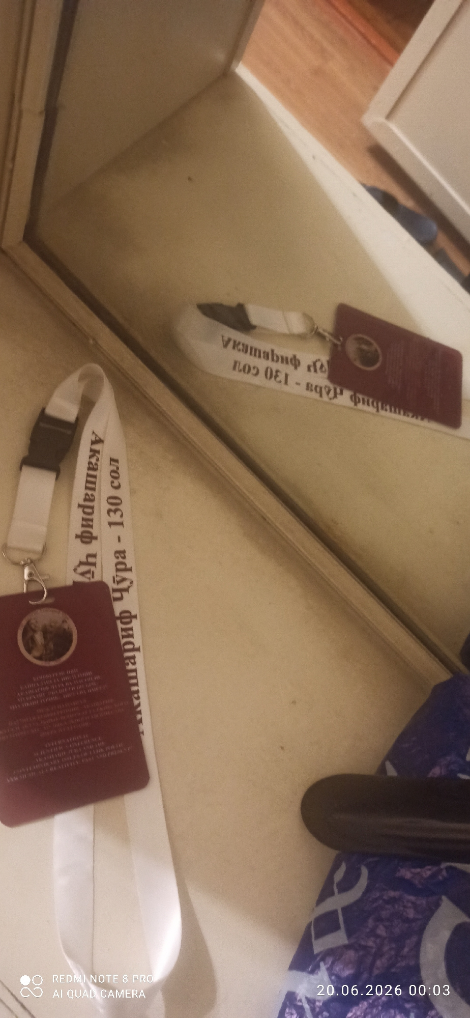 A lanyard with a maroon badge hangs on a plain, light-colored surface, likely a table or shelf. The badge has a circular emblem with what appears to be a portrait inside, and text in a smaller font below it. The lanyard itself is white with black text printed on it, in what appears to be Cyrillic script, reading "Акашшриф Чўра - 130 сол". Another lanyard, also white with black text, is visible in the reflection of a mirror, showing the words "Vкаmаbшf rlo3 OEI - Bqpş фицmaKIK". The mirror is positioned above the surface. The background is indistinct, but hints of wooden flooring and a white cabinet are visible in the reflection. The overall lighting is dim.