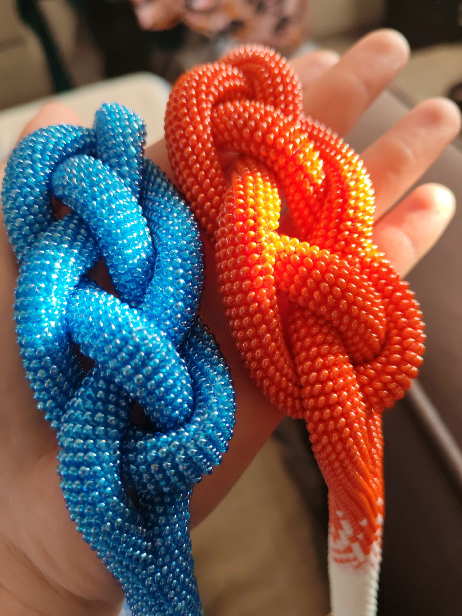 In a close-up, top-down shot, two hands are holding beaded objects. The object on the left is a thick, knotted rope made of small, shiny blue beads. The object on the right is a similar rope made of vibrant orange beads, transitioning to white at the bottom. The background is blurred, showing a soft, warm-toned surface that could be upholstery. The lighting creates a glossy effect on the beads, highlighting their texture and shimmer.