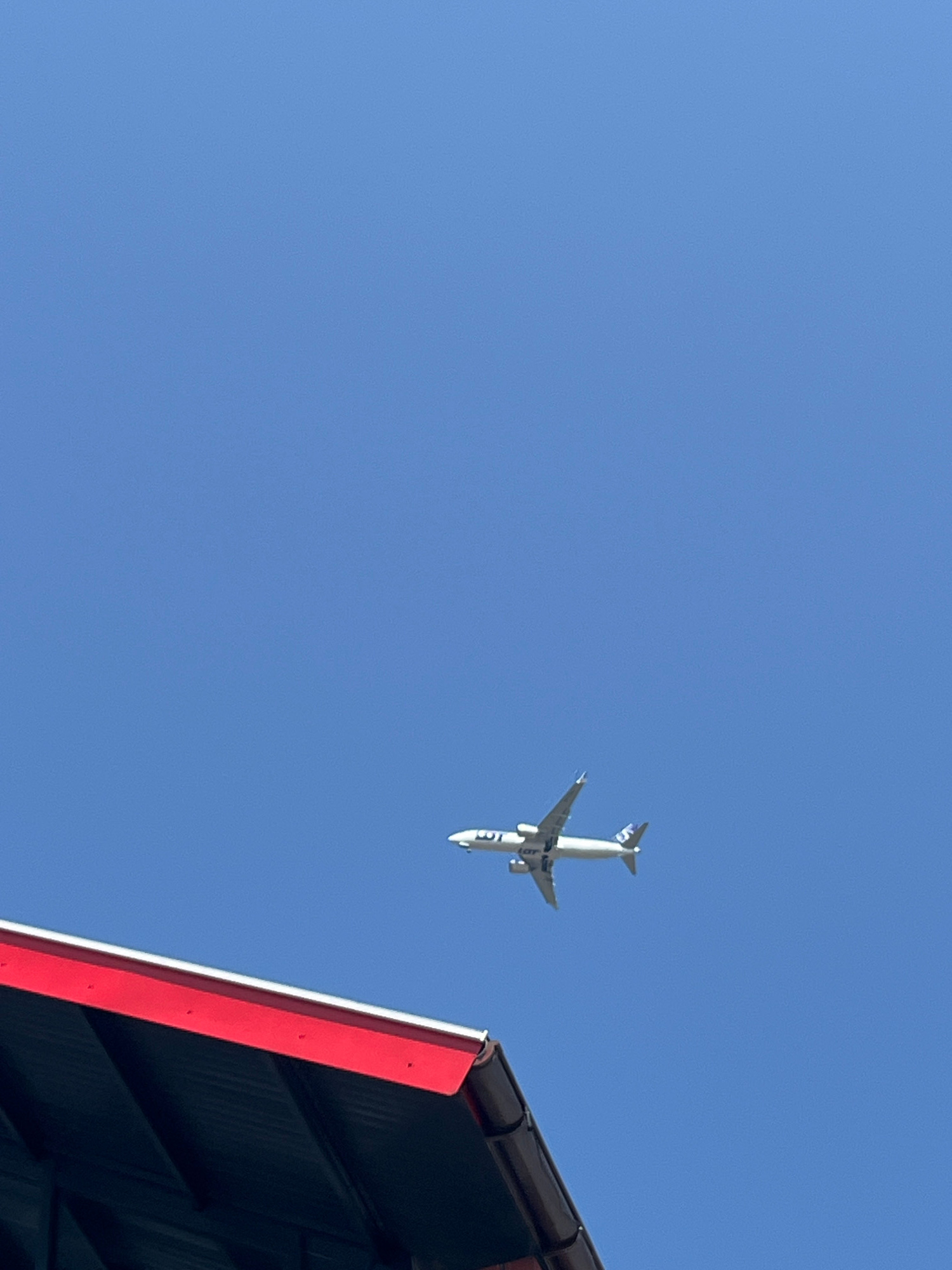 This image captures an airplane in flight against a clear blue sky. The airplane is white with blue accents and appears to have "LOT" written on its side. It is seen from below and to the side, with its wings outstretched. In the foreground, the edge of a dark-colored roof with a bright red trim is visible, partially obscuring the bottom right of the frame.
The scene suggests a daytime setting, likely sunny given the clarity of the sky and the bright colors. The presence of the airplane and the roof indicate an urban or suburban environment, possibly near an airport or under a flight path. The angle of the sun suggests it could be mid-morning or afternoon. No people are visible in the image. The overall mood is neutral, focusing on the contrast between the man-made structure and the aircraft against the natural backdrop of the sky. The crops highlight the airplane and the roof structure.