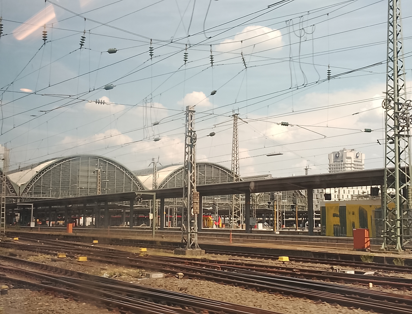 The image depicts a train station with multiple tracks and platforms under a large, arched, glass-covered roof. Overhead, a complex network of electrical wires and supporting structures crisscrosses the sky, indicating an active railway line.
In the foreground, numerous train tracks stretch into the distance, suggesting a busy transportation hub. Yellow markers are visible on the tracks, likely indicating points of interest or safety zones. The platforms are covered by dark awnings supported by steel columns, providing shelter for passengers. Some figures, appearing as small, indistinct shapes, are visible on the platforms, suggesting the presence of people, but their activities are not discernible.
Further in the background, a tall, modern building with many windows is visible above the station's roofline. To the right of the station, a bright yellow structure, possibly a utility building or maintenance area, stands prominently. A single red object, perhaps a bin or a marker, is positioned in front of it.
The sky is partly cloudy, with scattered white clouds against a light blue backdrop. The lighting suggests it is daytime, possibly late morning or early afternoon, as there are no strong shadows. The overall impression is that of a functional and active train station on a pleasant day. No visible text is identifiable in the image.