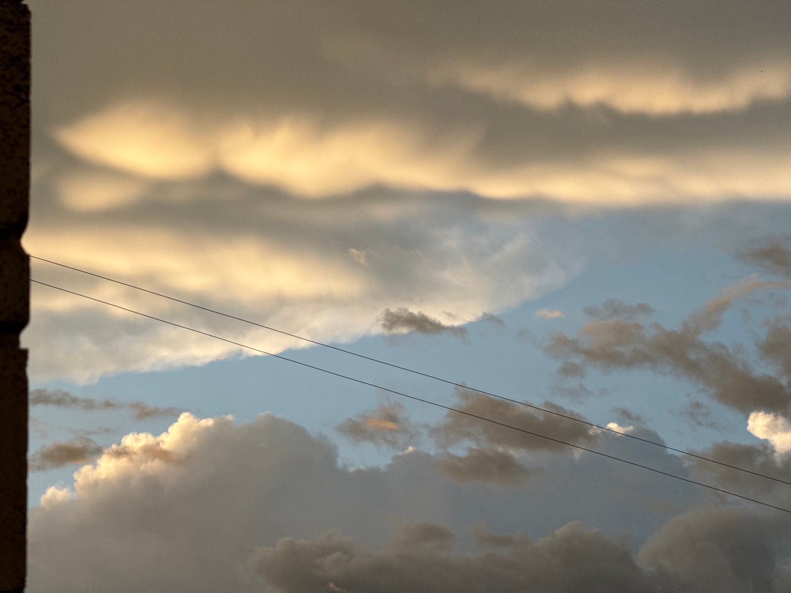 A close-up, low-angle shot captures a sky filled with dramatic clouds at what appears to be dusk. The left side of the frame is dominated by a dark, textured vertical element, possibly a building or pillar, encroaching into the scene. A band of bright, golden-yellow light illuminates a thick layer of clouds directly above, with darker, more diffused clouds forming distinct bands above and below this illuminated layer.
Further down, a bright blue sky is visible through a gap in the clouds, with scattered cumulus clouds. Two thin, dark power lines crisscross the frame from the upper left to the lower right, cutting through the cloud formations and the blue sky. The overall impression is one of atmospheric turbulence and changing light conditions.