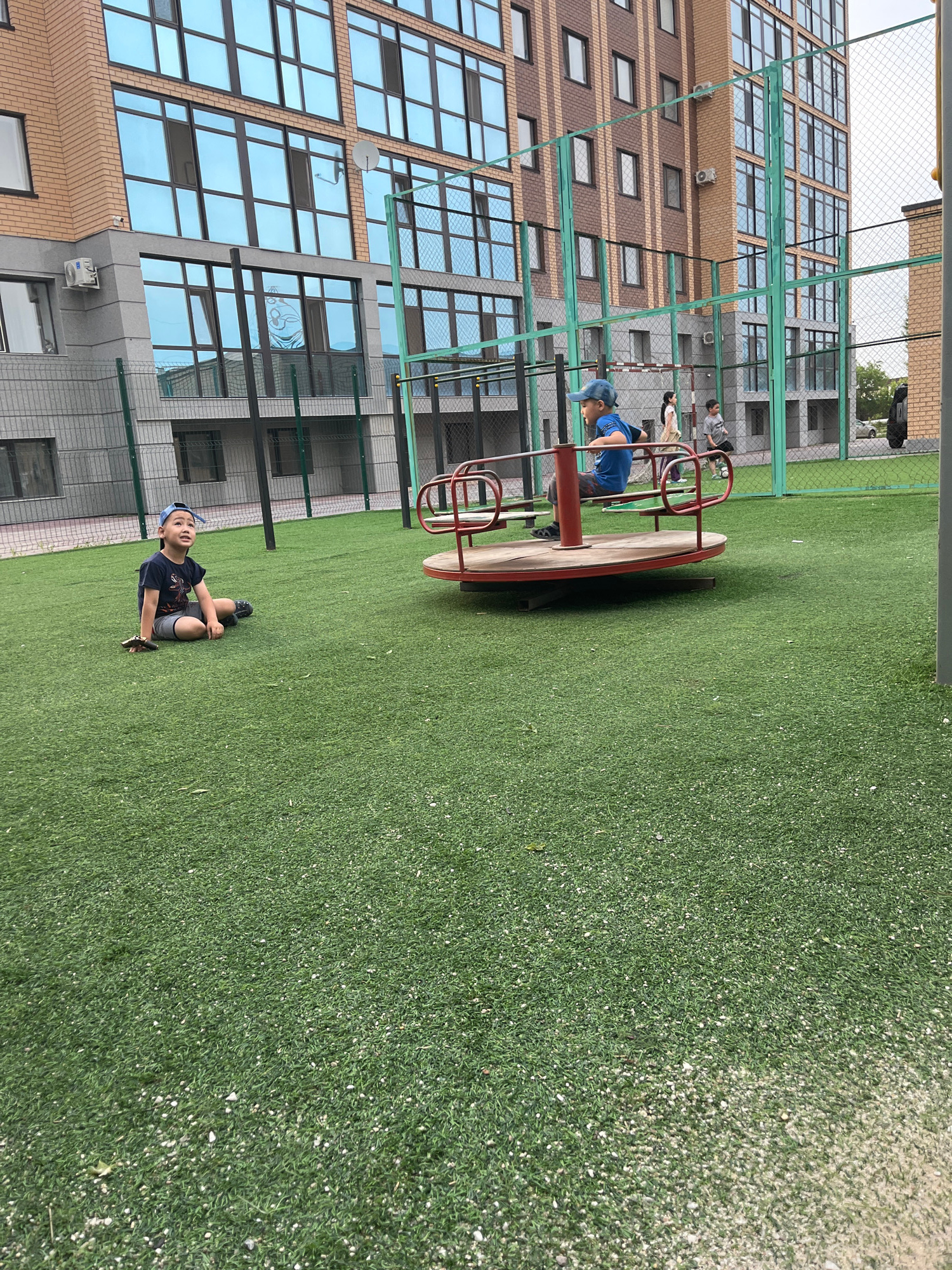 This image depicts a playground with artificial turf flooring located in Kokshetau, Kazakhstan. In the foreground, a young boy wearing a blue baseball cap and a dark t-shirt sits on the grass, looking upwards. To his right, another boy in a blue shirt is seated on a red merry-go-round. Behind them, two other children are visible, one standing and the other in motion, possibly playing. The background features a modern apartment building with large windows. The playground is enclosed by a green fence. The overall scene is of an outdoor recreational area.