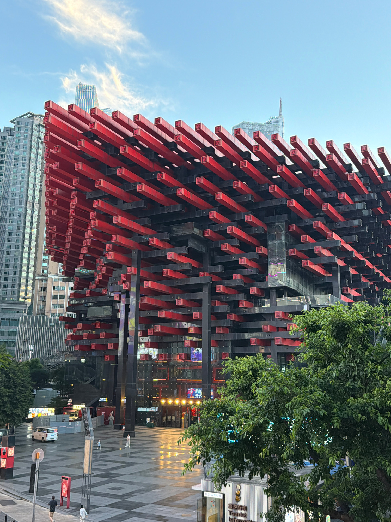 An exterior, medium-angle shot captures a striking modern building with a facade of protruding red rectangular elements, some displaying red square symbols. The building is surrounded by other high-rise structures and is situated in an urban plaza with a patterned paving design. A lush green tree dominates the lower right portion of the frame, partially obscuring the view of the building's lower levels. In the plaza, several people are visible, along with a white van and street furniture. The sky above is a clear blue with wispy white clouds. This scene is located in Yuzhong District, China.