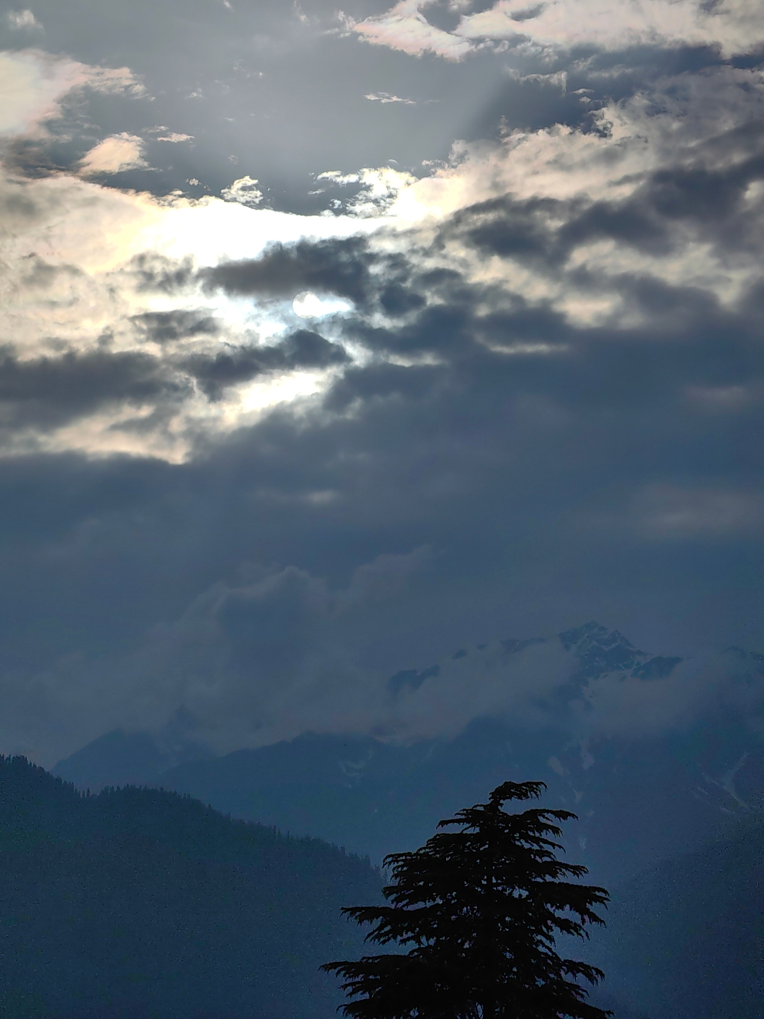 This image captures a dramatic sky over a mountainous landscape in Siumsa, India. The scene is dominated by a turbulent sky filled with dark, heavy clouds interspersed with brighter, sunlit patches. Rays of sunlight pierce through the clouds, creating a dynamic and almost ethereal effect. The sun itself is partially obscured by the clouds, casting a soft, diffused glow.
Below the dramatic sky, layers of mountains recede into the distance. The closest mountain range is densely forested with dark, silhouetted trees, creating a stark contrast with the brighter sky. Further in the background, snow-capped peaks are visible, peeking through thinner clouds, hinting at a high-altitude location. In the foreground, the silhouette of a large, coniferous tree is prominent on the right side of the frame, its branches reaching upwards into the cloudy sky.
The overall mood is one of awe and perhaps a touch of melancholy, evoked by the powerful play of light and shadow. The weather appears to be changing, possibly indicating an approaching storm or the aftermath of one. The time of day is difficult to determine precisely due to the cloud cover, but the quality of light suggests it could be late afternoon or early morning, when the sun is lower in the sky. There is no visible text in the image.