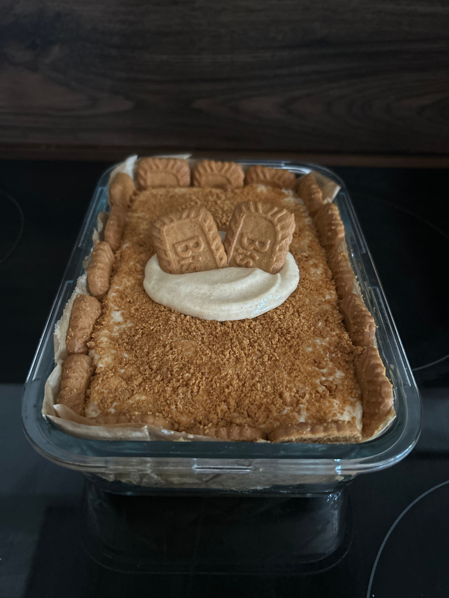 A rectangular glass baking dish filled with a dessert is centered in the frame. The dessert appears to be a no-bake cheesecake or similar layered confection, with a base of crushed biscuits and a creamy filling. The top surface is covered in finely crumbled biscuits, with a dollop of lighter cream and two embossed biscuits placed in the center. The sides of the dish are lined with whole biscuits, and a layer of parchment paper is visible beneath the dessert. The dish rests on a dark, reflective surface, possibly a stovetop, with a wooden textured background above.