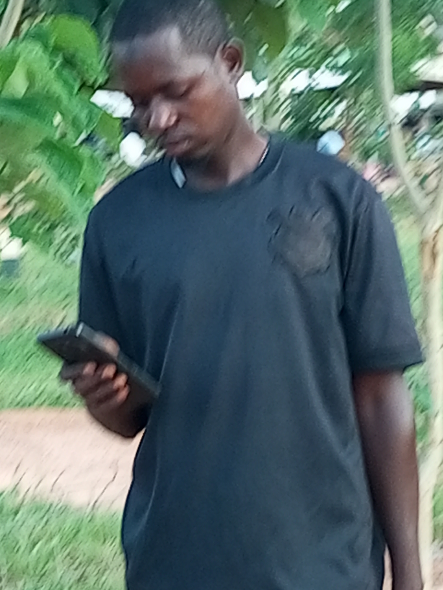 A medium shot captures a Black man from the chest up, looking down at a dark rectangular object, likely a smartphone, held in his left hand. He is wearing a dark short-sleeved t-shirt with a textured, dark emblem on the left chest area. The background consists of out-of-focus greenery, including leaves and grass, suggesting an outdoor setting. A hint of a light-colored pathway is visible in the lower left quadrant. The overall lighting suggests it is daytime. There are no other people or identifiable structures in the immediate vicinity. The environment appears to be a park or a natural setting within the city of Gombe, Nigeria.