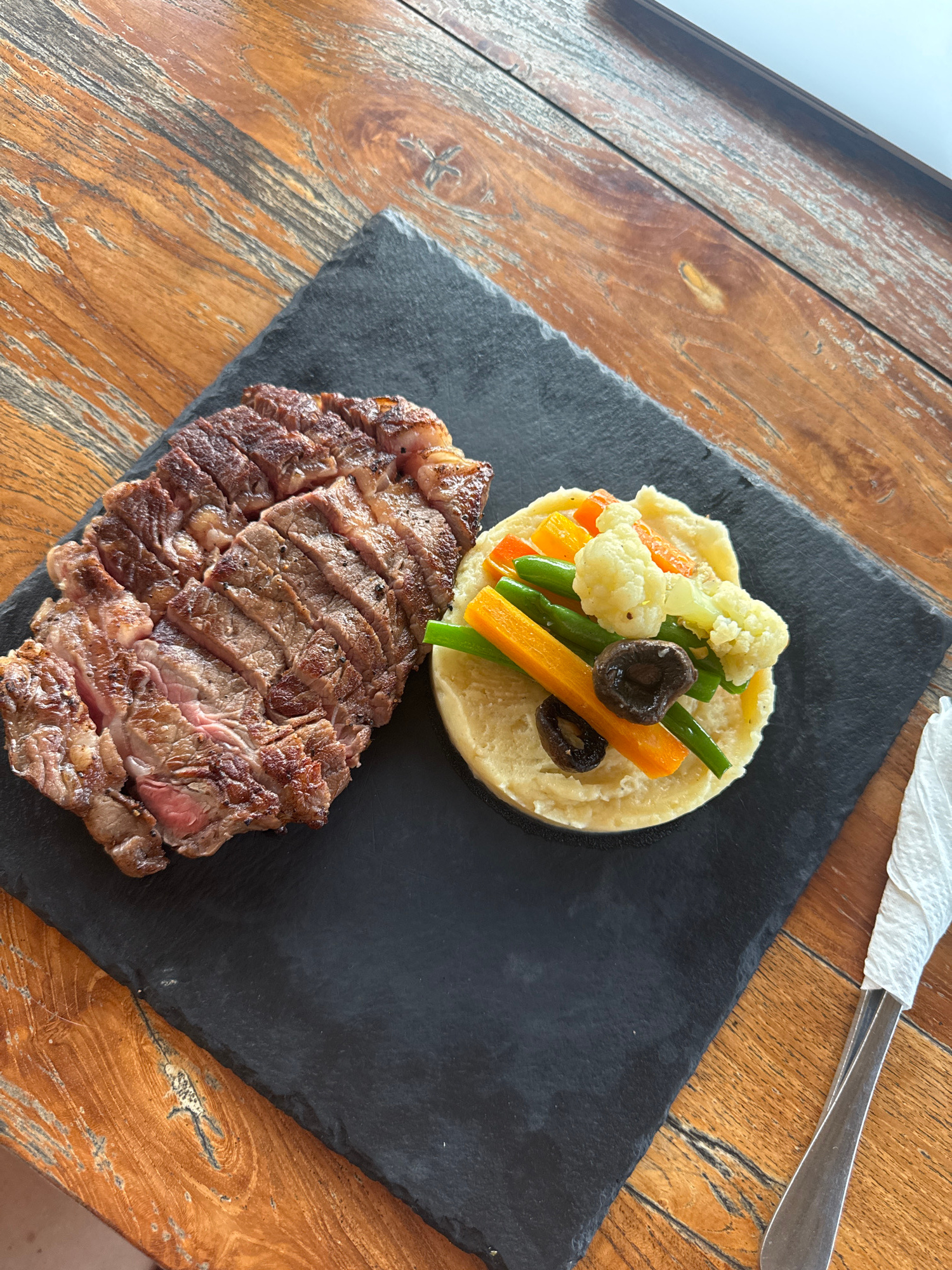A top-down, close-up shot features a sliced steak and mashed potatoes with vegetables on a black slate plate, which is placed on a rustic wooden table. The steak, cooked to a medium-rare, is sliced and seasoned with black pepper. Accompanying the steak is a mound of creamy mashed potatoes topped with blanched cauliflower florets, green beans, carrot sticks, and two slices of black olives. A silver fork, partially wrapped in a white napkin, rests on the right side of the frame, also on the wooden table. The wooden table has a prominent grain pattern and appears to be outdoors, suggesting a restaurant setting in Nusa Penida, Indonesia.
