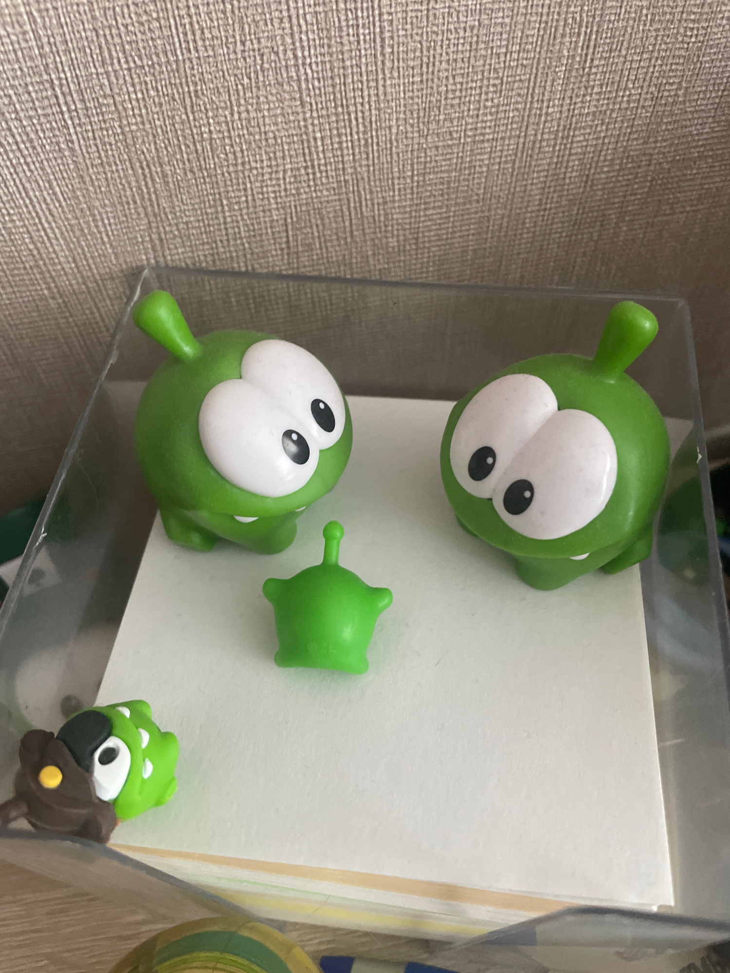 The image shows a collection of small, green, cartoon-like plastic toys, resembling characters from the game "Cut the Rope." Two larger figures, each with large white eyes and small black pupils, are positioned on a white surface. A smaller, green alien-like figure with a pointed head and a wider base stands between them. In the lower left corner, a brown and green toy with large eyes and a yellow circular detail is visible. The toys are contained within a clear plastic box. The background appears to be a textured, light brown wall. There is no indication of the location being Moscow, Russia.