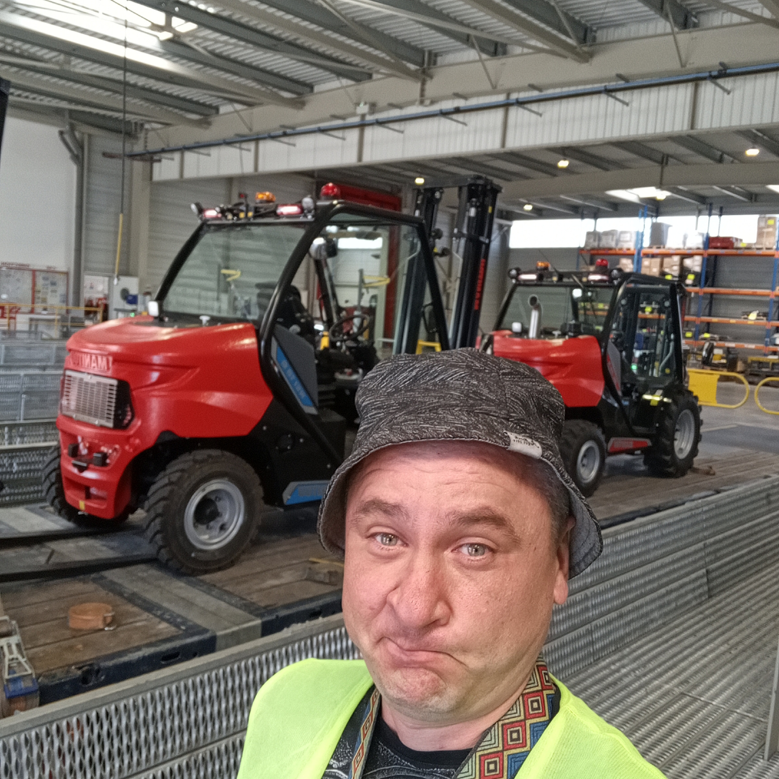 A man with light skin, brown eyes, and short brown hair wears a grey patterned bucket hat and a neon yellow high-visibility vest over a dark t-shirt with a geometric pattern around the neckline. He makes a pouty facial expression. He is standing in a warehouse or industrial setting. Behind him, to the left, is a red and black forklift. To the right of the red forklift, and further back, is another black forklift. Both forklifts appear to be on a flatbed trailer. The forklifts have safety lights on top. The warehouse has a metal ceiling with exposed beams and fluorescent lighting. Shelving units with boxes are visible in the background on the right. The floor appears to be concrete and metal grating. The man's posture suggests he is taking a selfie. The overall scene is brightly lit, indicating daytime. The location is identified as Ancenis, France.