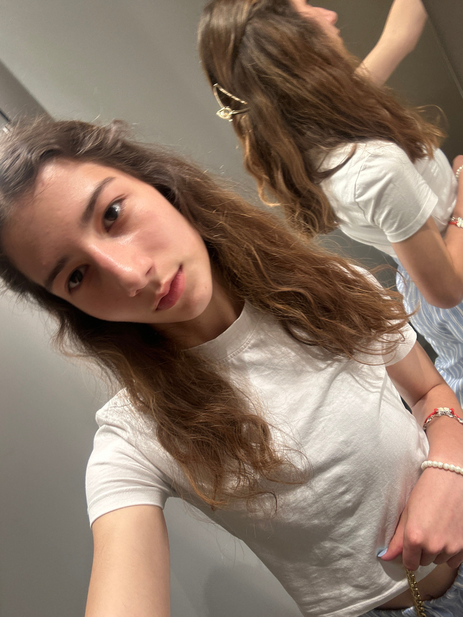 A young person with long, brown, wavy hair is taking a selfie in a fitting room. They are wearing a white t-shirt. The person is positioned close to the camera, with their face slightly tilted. In the background, a reflection shows another person, also with brown hair, wearing a white t-shirt and striped pants. The fitting room appears to have gray walls. The image does not provide any specific visual cues to pinpoint its location within Ruse, Bulgaria.