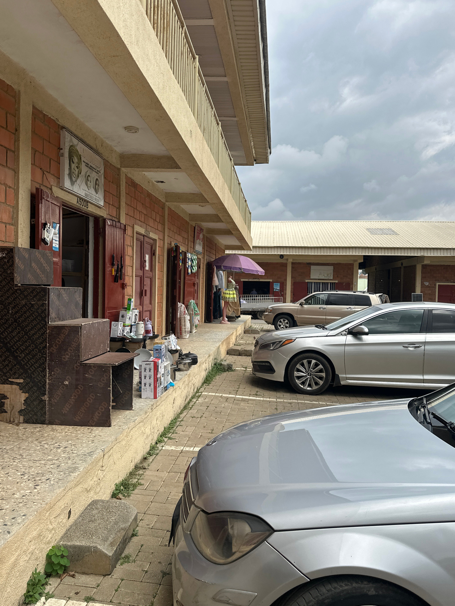 This image captures a street scene outside a row of shops in Abuja, Nigeria. The shops are constructed with red bricks and have a covered walkway in front, supported by a light-colored railing. Some shops display goods on the ground, including boxes, kitchenware, and packaged items.
A woman in traditional attire, holding a purple umbrella, stands near one of the shops. Several vehicles are parked in front of the shops, including a silver sedan and a beige SUV. The ground is paved with interlocking bricks. The sky is overcast with gray clouds. The scene appears to be a commercial area in Abuja, likely a market or a strip mall.