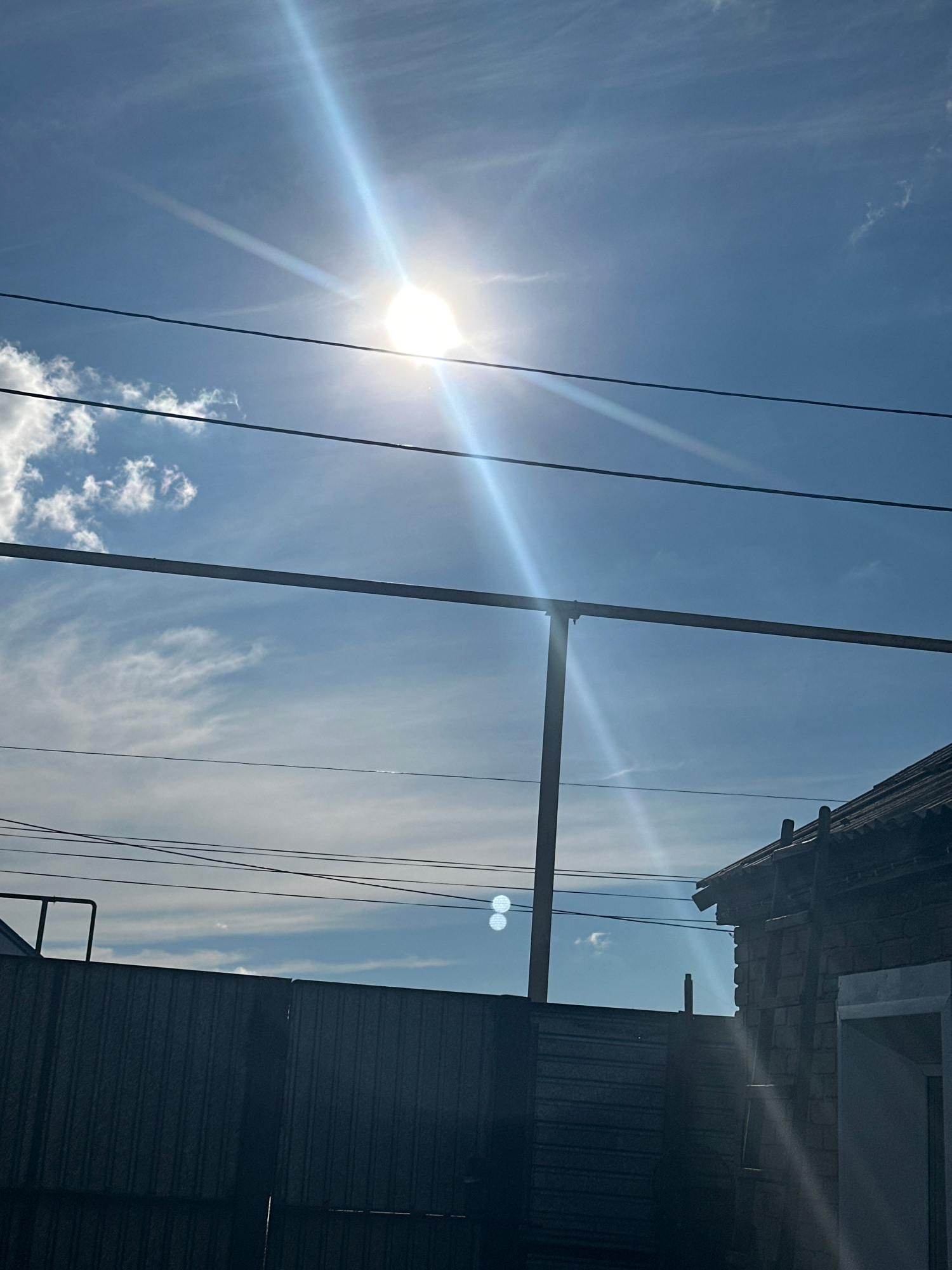 The image depicts an outdoor scene with a clear blue sky, some wispy white clouds, and the sun shining brightly, creating lens flares. Several dark power lines are visible, running horizontally across the frame. In the upper right portion of the image, a section of a building is visible, with a dark, corrugated metal wall and a roof that appears to be made of tiles. There are also hints of a window and possibly a gutter system on the building. A vertical pole, likely a utility pole or part of a structure, intersects the power lines. The overall impression is of a sunny day, possibly midday, with a utilitarian or semi-industrial setting. The perspective is looking upwards towards the sky and the building. There is no visible text in the image.