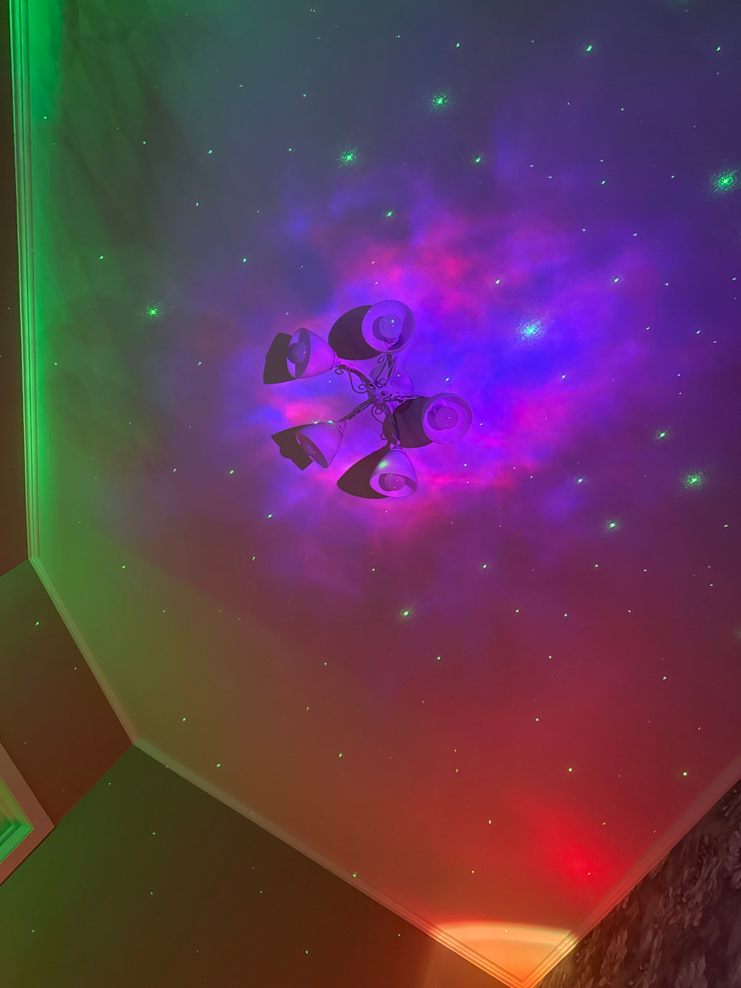 The image shows a ceiling decorated with a galaxy projector. The projector creates a nebula effect with purple and pink hues, along with scattered green stars. A ceiling fan with multiple light fixtures is partially visible in the upper center of the image. The ceiling is lit with a gradient of colors, transitioning from green on the left to purple in the center and red/orange on the right. The lighting is dim and atmospheric. There are no people or animals present in the scene. The location is described as Unknown, Unknown.