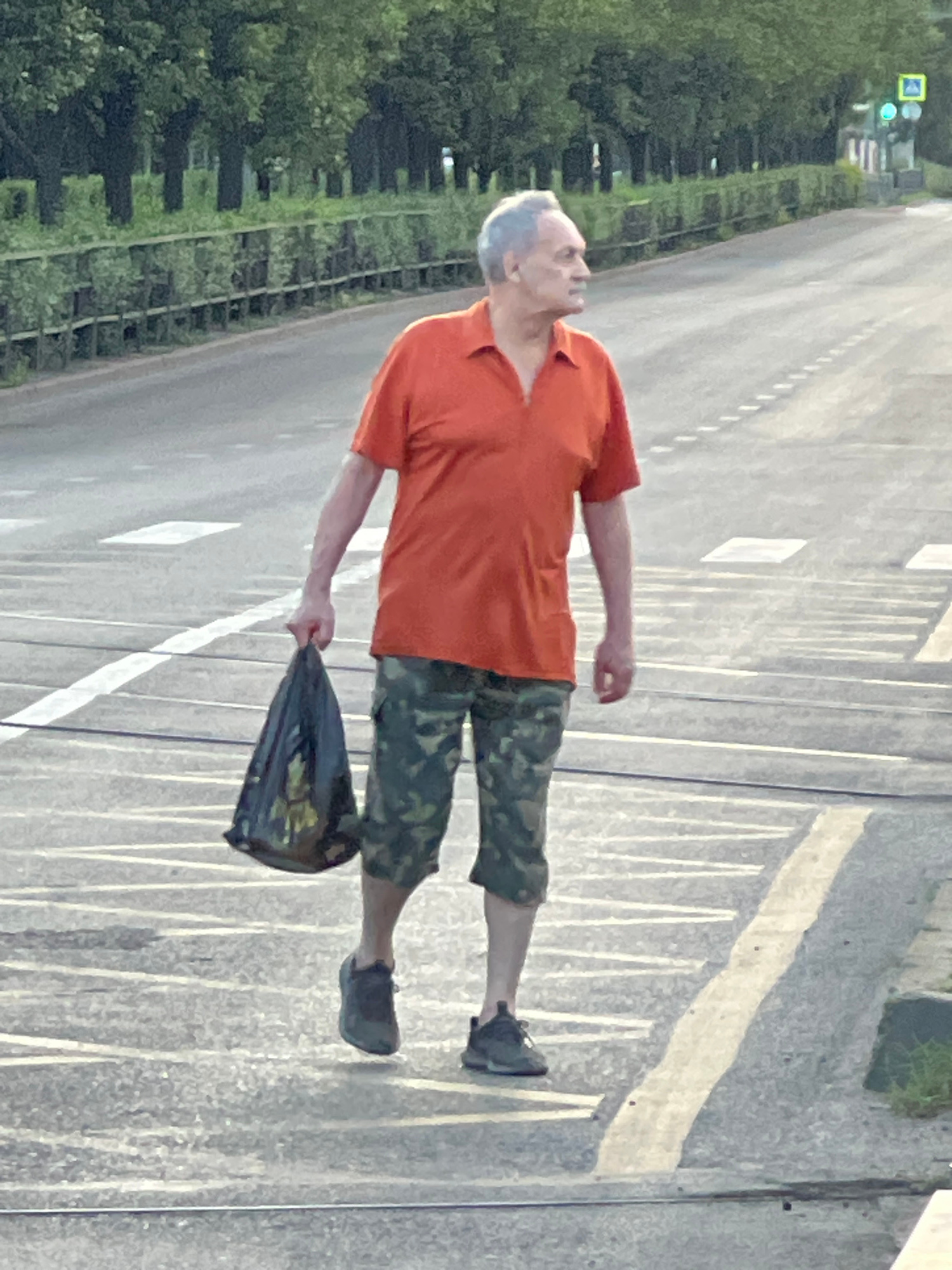 An elderly man with graying hair is walking across a crosswalk, carrying a black plastic bag in his left hand. He is wearing an orange polo shirt and camouflage shorts. The man is looking to his right. The crosswalk is on a paved road with white dashed lines indicating lanes. In the background, there are trees and a traffic light. The scene appears to be in an urban environment in Krasnodar, Russia.